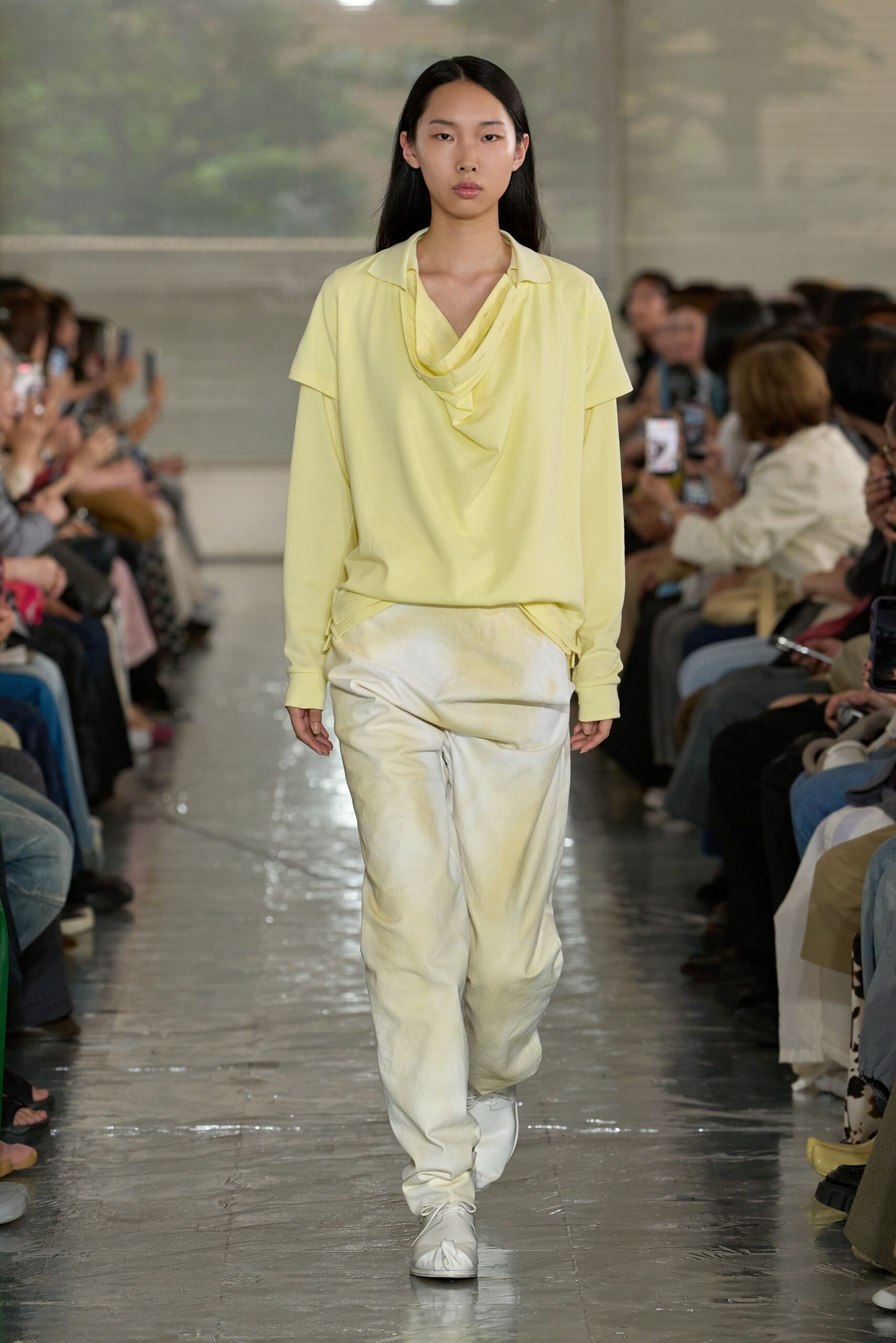 Butter-yellow draped top with a soft cowl neck layered over long jersey sleeves and a tiny notch collar. Ivory washed-satin trousers, gently puddled at the ankle. White gathered slippers. Early check-in, hotel corridor still smelling of bleach.