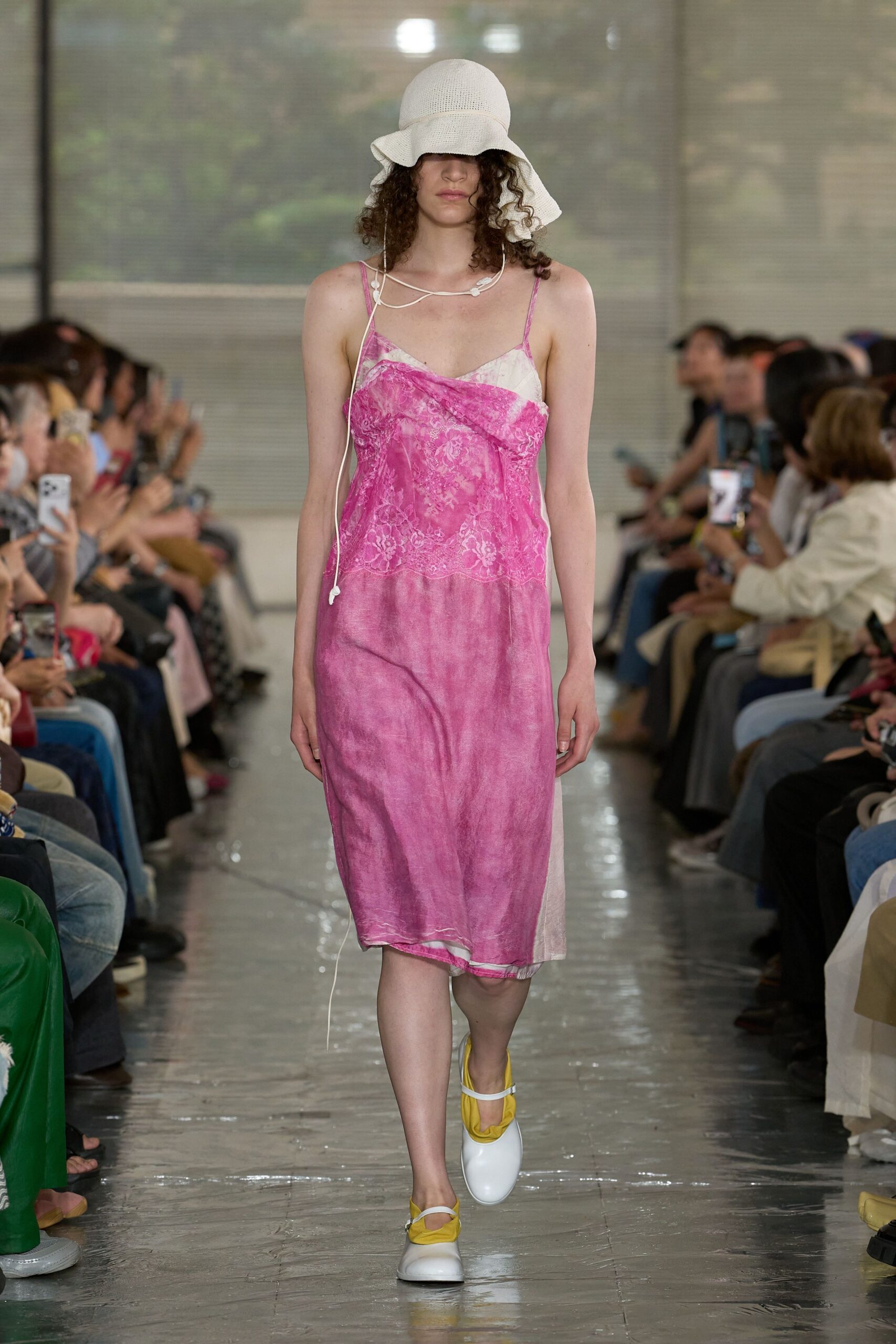 White open-weave sunhat with a drooping brim, pink slip dress washed from blush to fuchsia with lace laid across the bust, white cord looped loose at the neck, white Mary Janes with citron collars. After swimming, tiled lobby still damp.