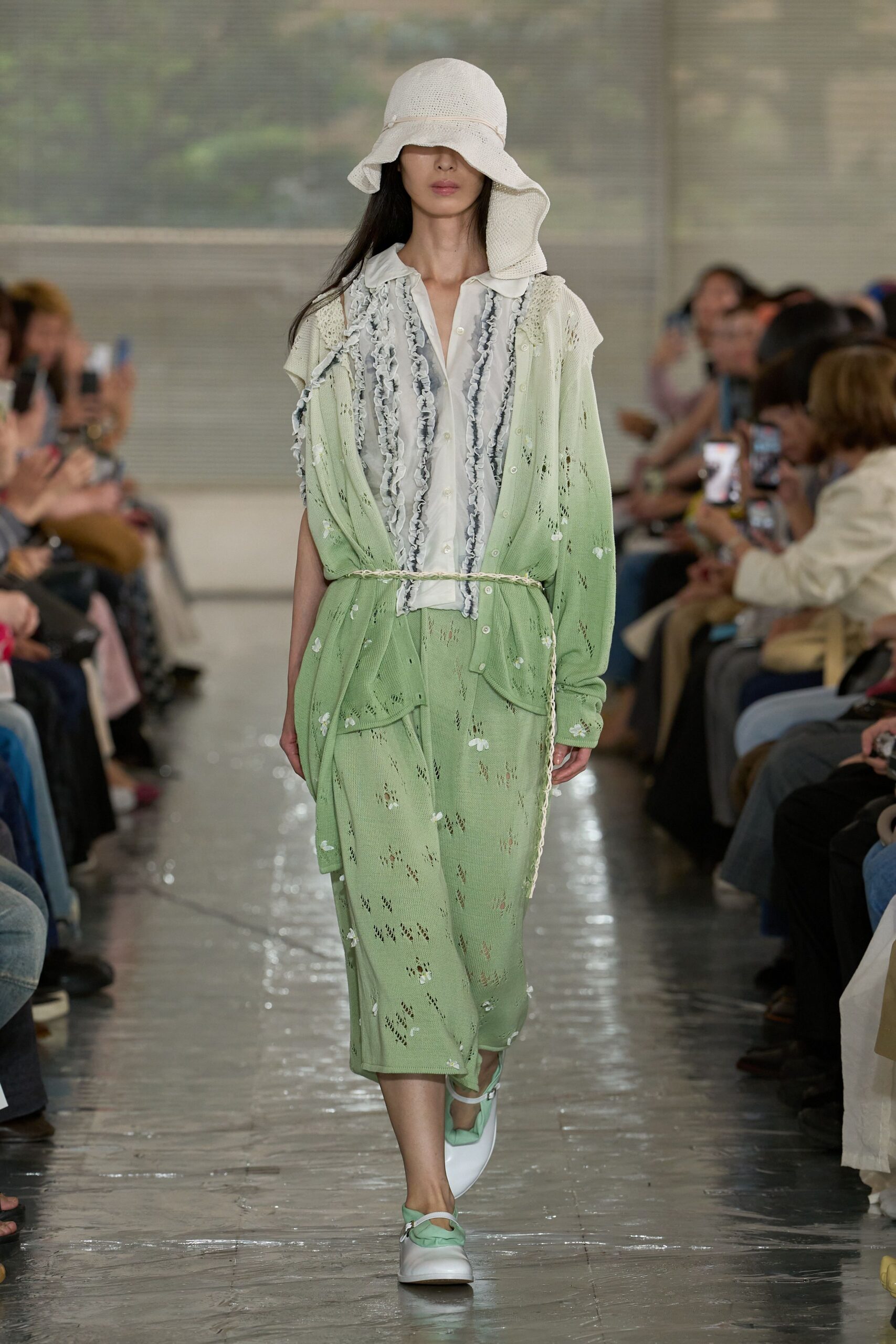 Floppy white open-weave hat, sheer white shirt trimmed with pale grey ruffles, and a pistachio pointelle knit cardigan and midi skirt scattered with tiny appliqué blossoms, tied with a twisted cord belt. Greenhouse café, rain still on the glass.