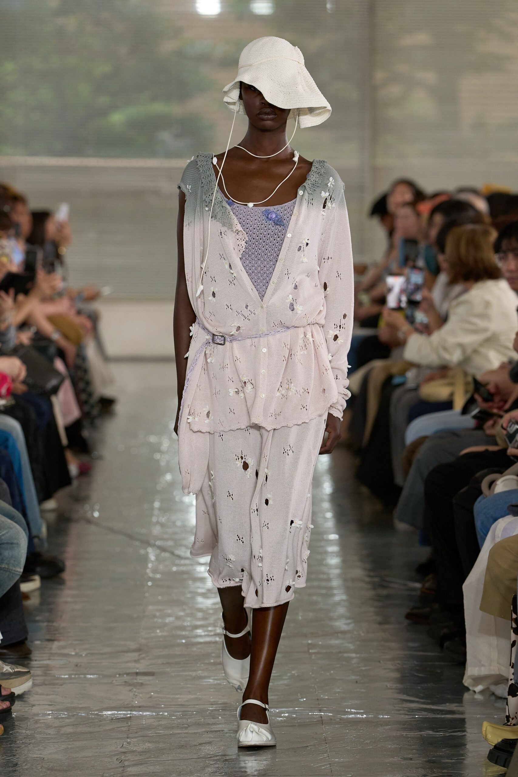 White woven sunhat with a drooping brim and chin cords. Blush pointelle cardigan scattered with tiny appliqué flowers over a lilac open-knit cami, tied with a narrow belt. Matching skirt, white strap flats. After the hotel pool closes.