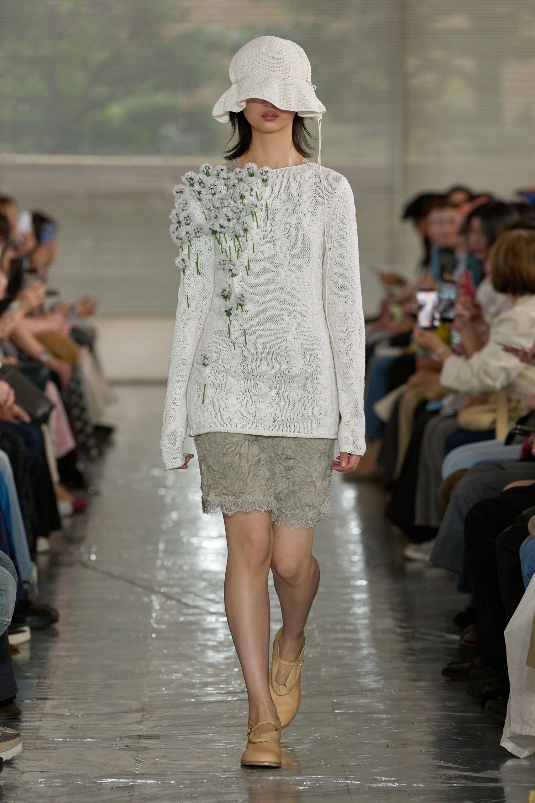 White open-knit cloche hat with a drooping brim. Ivory sweater dress in loose stockinette, one shoulder crowded with tiny tufted flowers on green stems. Crinkled putty slip hem, beige Mary Janes. Greenhouse aisle after the misting cycle.