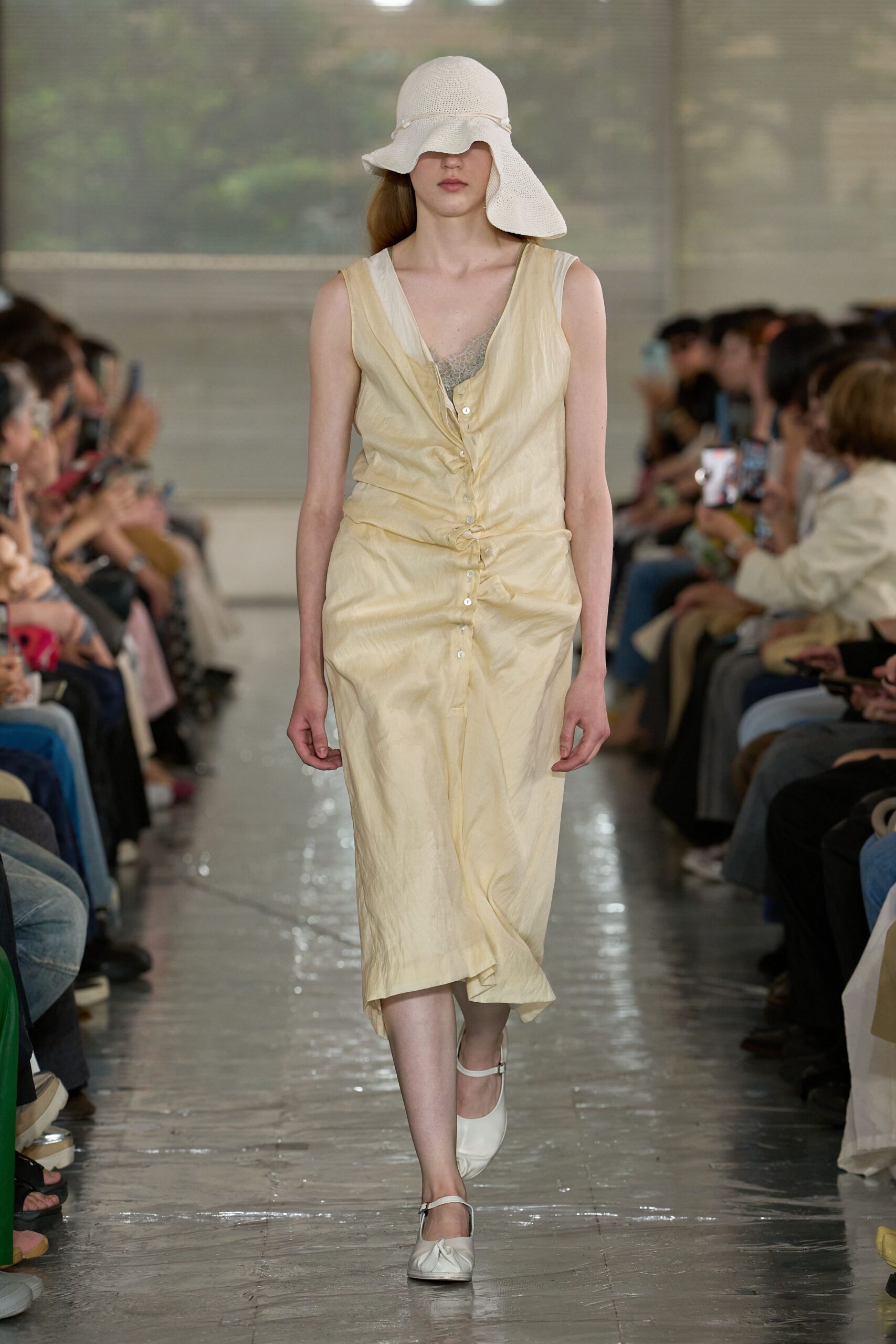 Buttercream sleeveless dress in crinkled satin, pearl buttons pulling the front into soft gathers. Sage lace camisole peeking at the V, white Mary Janes, floppy open-knit sun hat. Noon on a hotel terrace, tablecloths lifting in the heat.