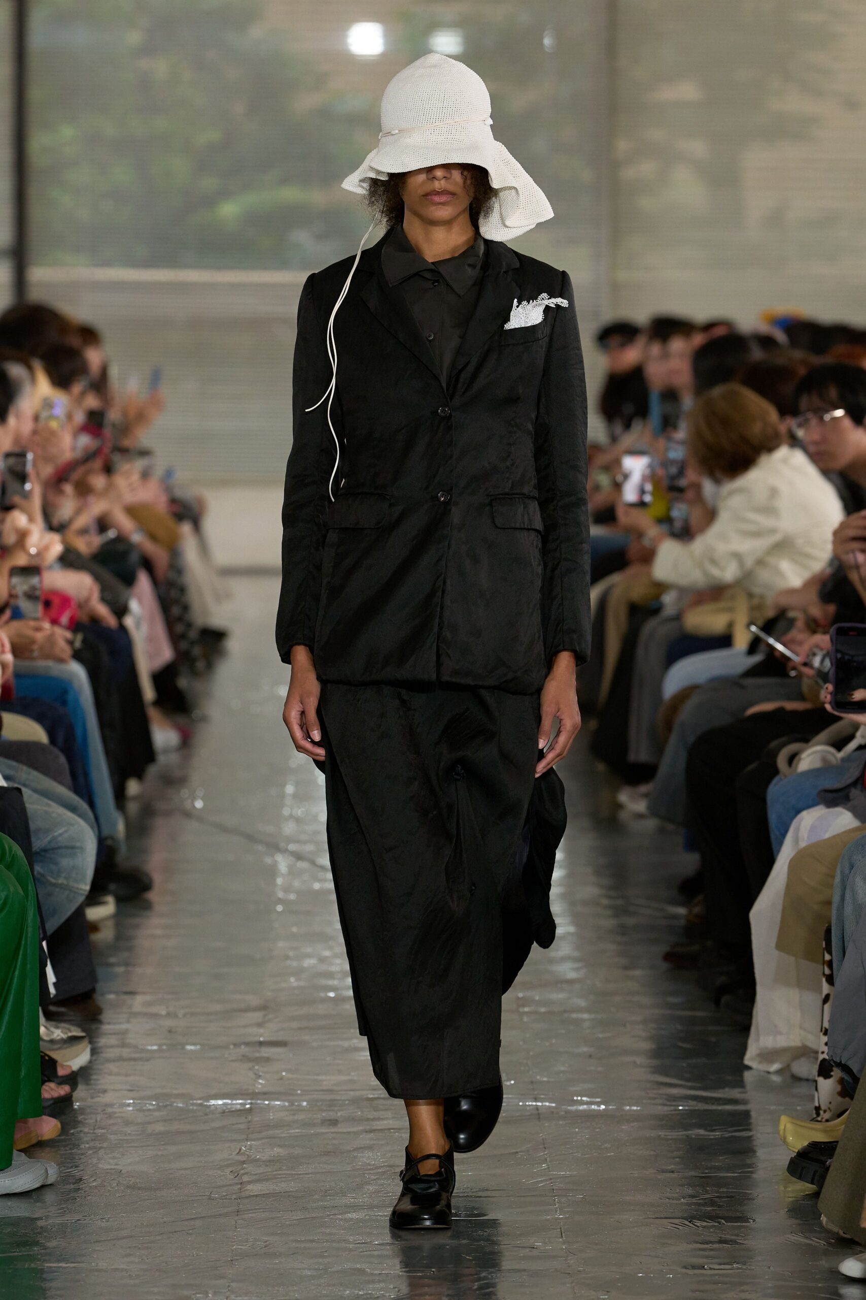 White open-weave sunhat with a floppy brim and trailing chin cord, black crinkled blazer over a sharp-collared shirt, long black skirt with a soft collapse at the shin, rounded black flats. Hotel lobby at noon, still damp from rain.