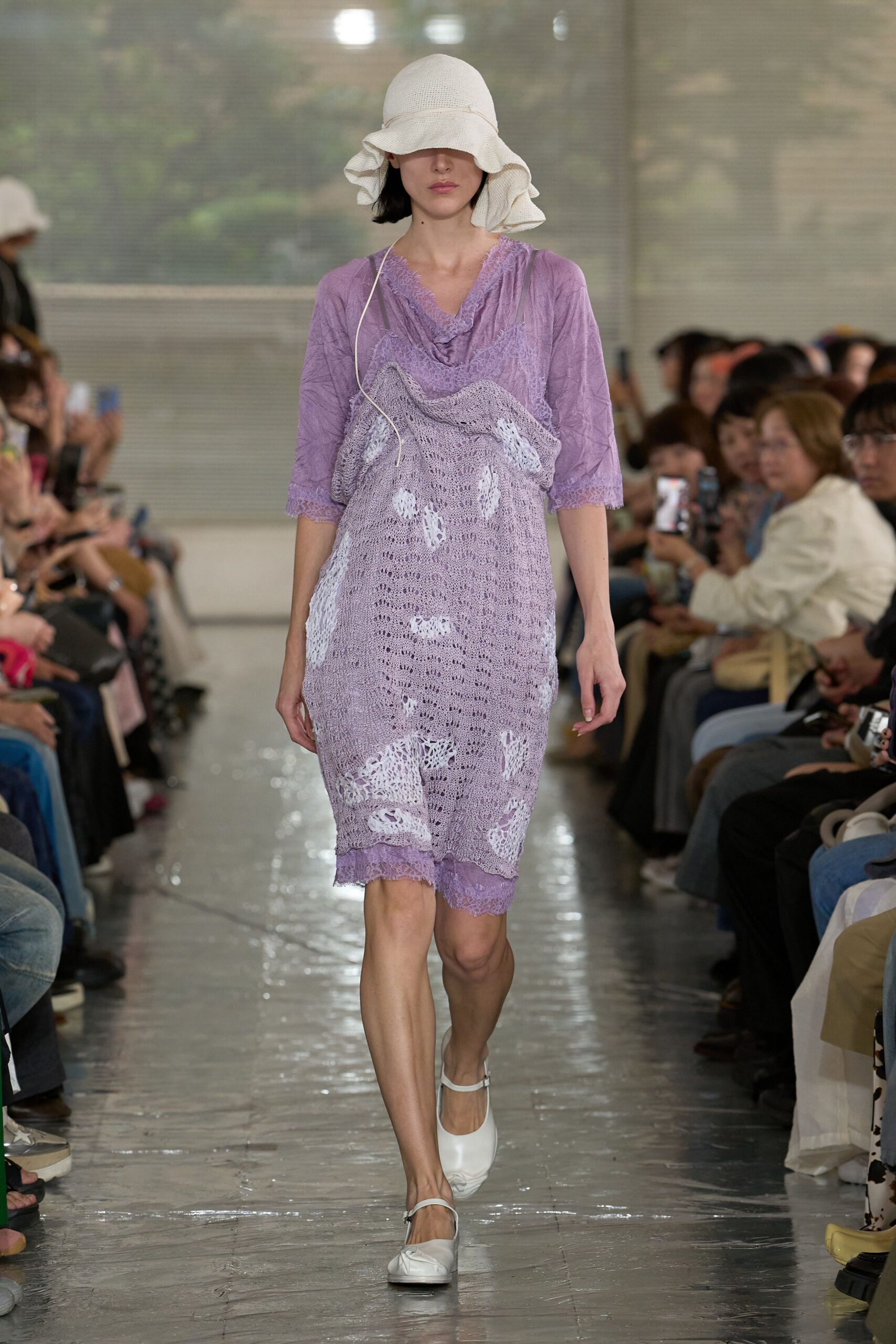 Cream open-weave sun hat with a ruffled brim. Crinkled lilac slip dress edged in eyelash lace, draped with a loose lavender crochet layer full of worn-through holes. White strap flats. Humid conservatory aisle, just after watering.