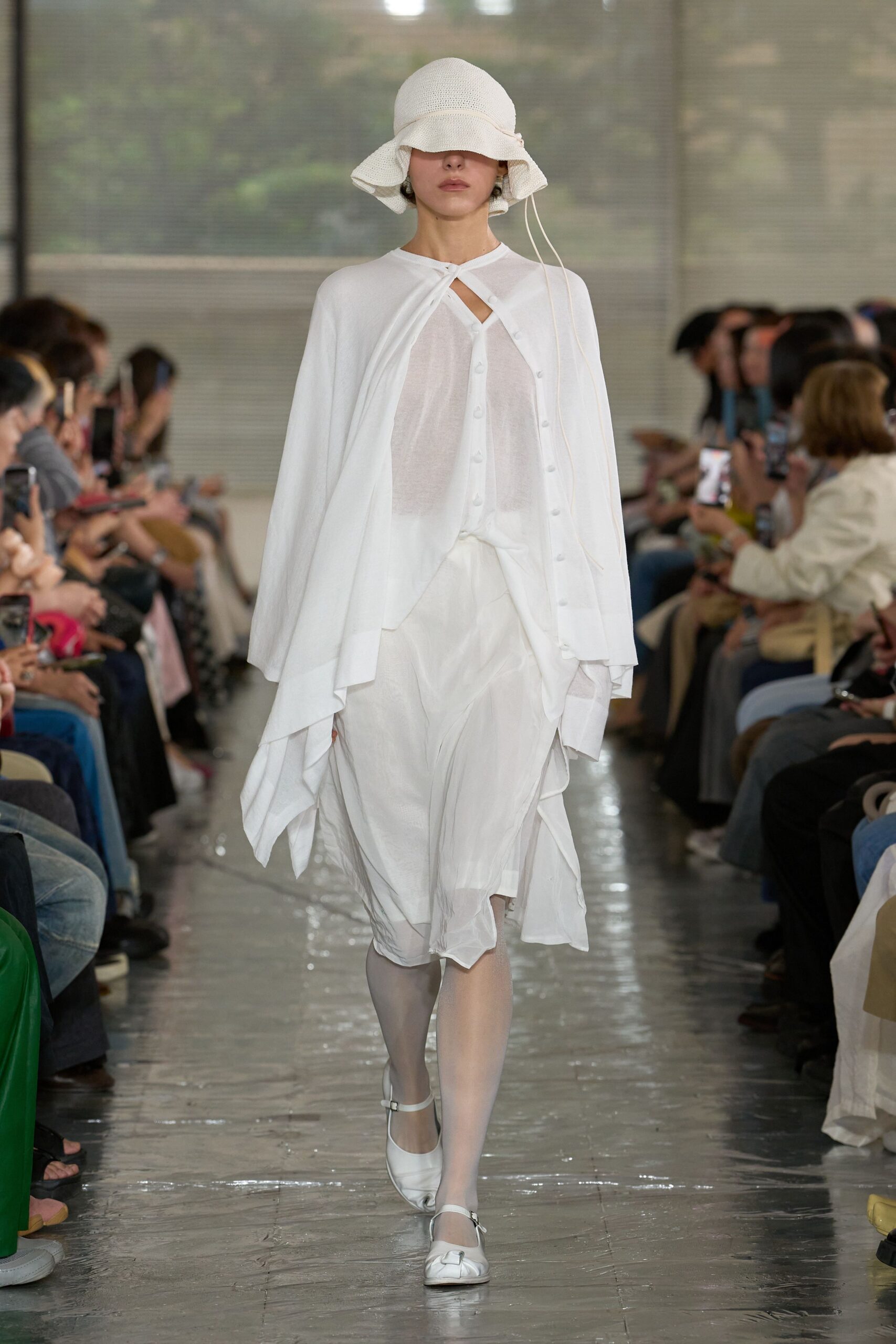 White openwork bucket hat with a floppy brim and dangling ties, sheer white capelet fastened at the throat over a gauzy button-front dress, white tights, strap flats with a square bow. Crossing a tiled conservatory after rain.