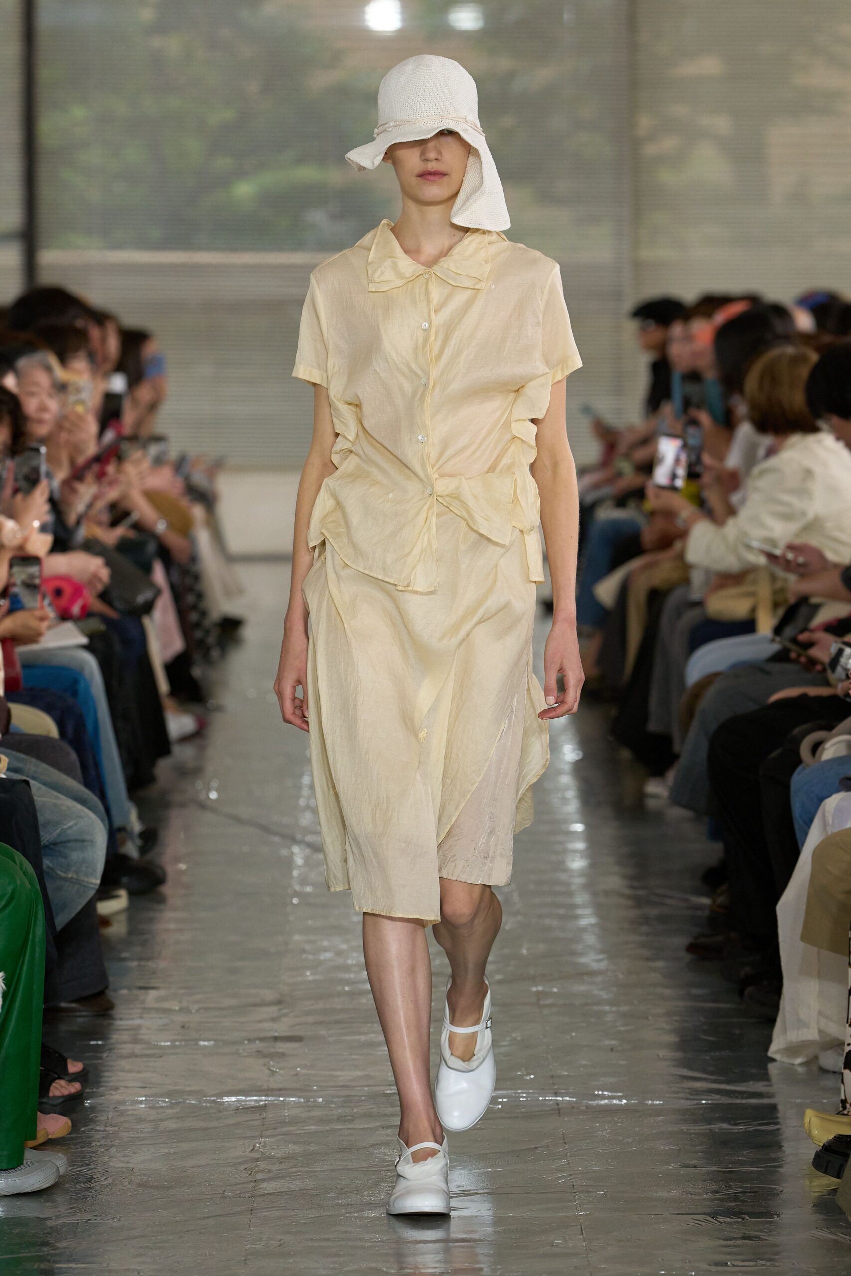 White open-weave cloche hat with a floppy side panel. Buttercream short-sleeve blouse in crinkled satin, ruched into soft knots at the waist. Matching wrap skirt, white Mary Janes. Humid conservatory aisle, just before the storm breaks.
