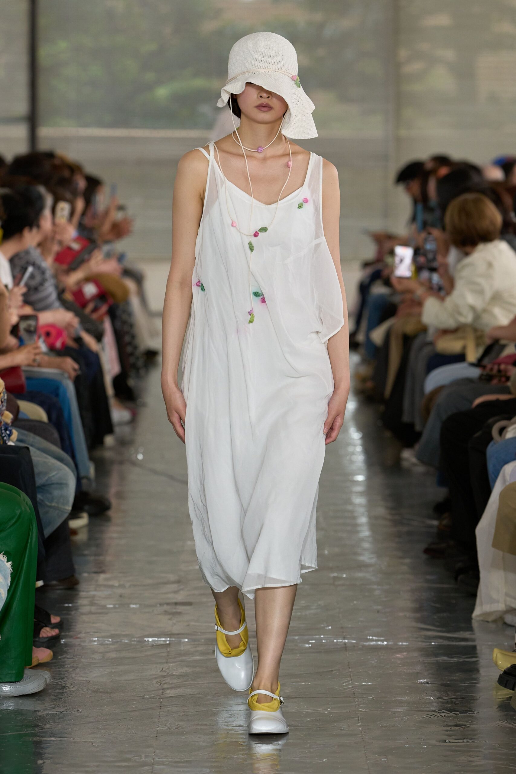 White open-weave cloche hat with a wavering brim, sheer white slip dress cut with a drooping patch pocket and scattered tiny rosettes, long cord necklace, white Mary Janes lined in citron. Greenhouse corridor after the watering.
