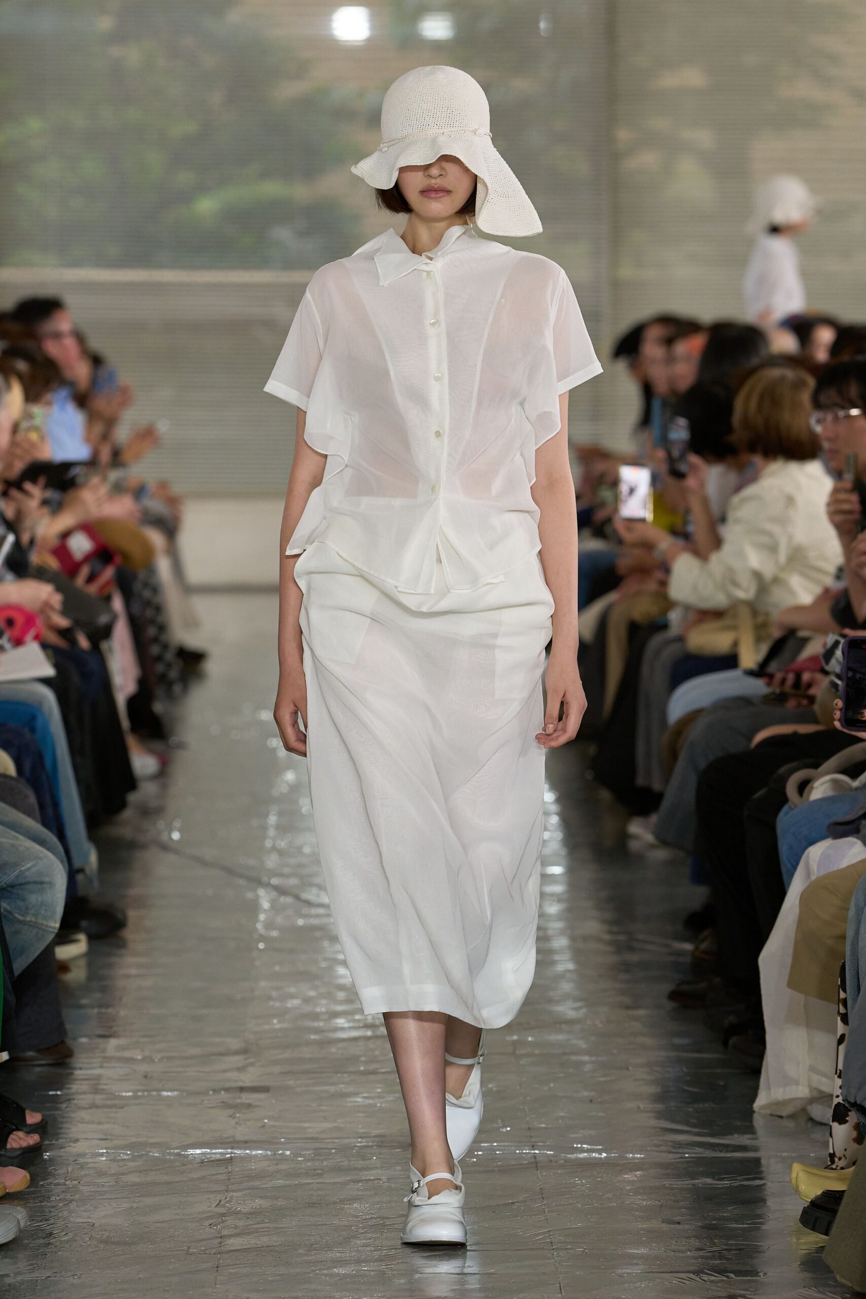 Open-knit white sun hat with a wavering brim. Sheer white blouse in crisp voile, cut with fluttering side panels and a tiny folded collar. White draped skirt, white strap shoes. Noon in a glasshouse after the watering cans.