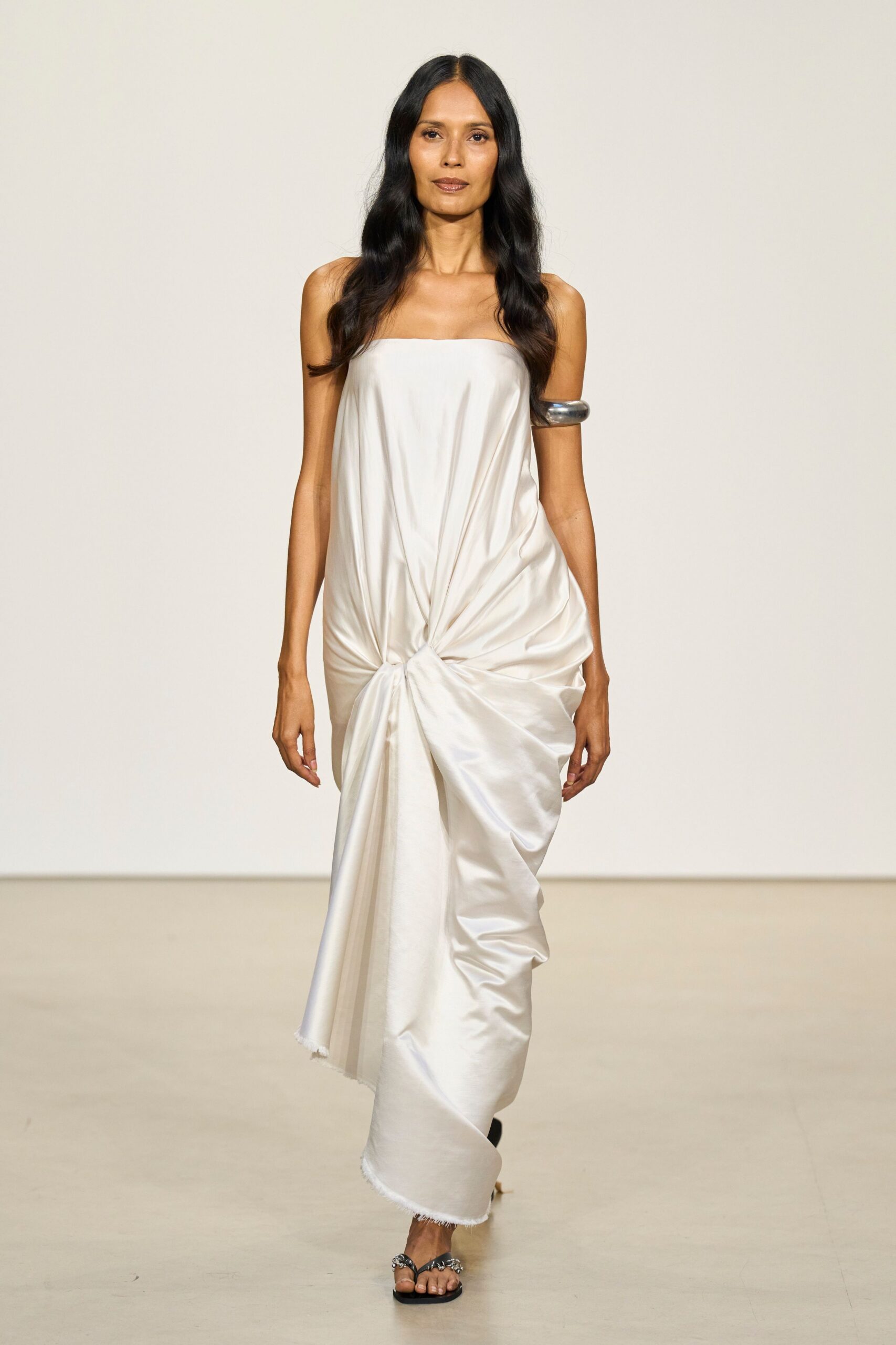 Ivory satin tube dress, twisted low at the hips into heavy drape. Chunky silver cuff gripping the upper arm. Black thong sandals with knotted silver over the toes. Courthouse steps after the ceremony, bouquet traded for someone's cigarettes.