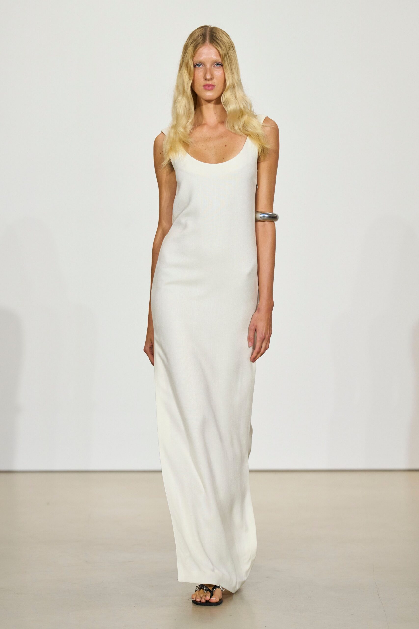 Ivory column slip dress in fluid jersey, scooped low at the chest. Oversized silver cuff hugging the upper arm. Black flat thong sandals with clustered metal at the toe. Walking barefoot-adjacent across a hotel terrace after the pool has closed.