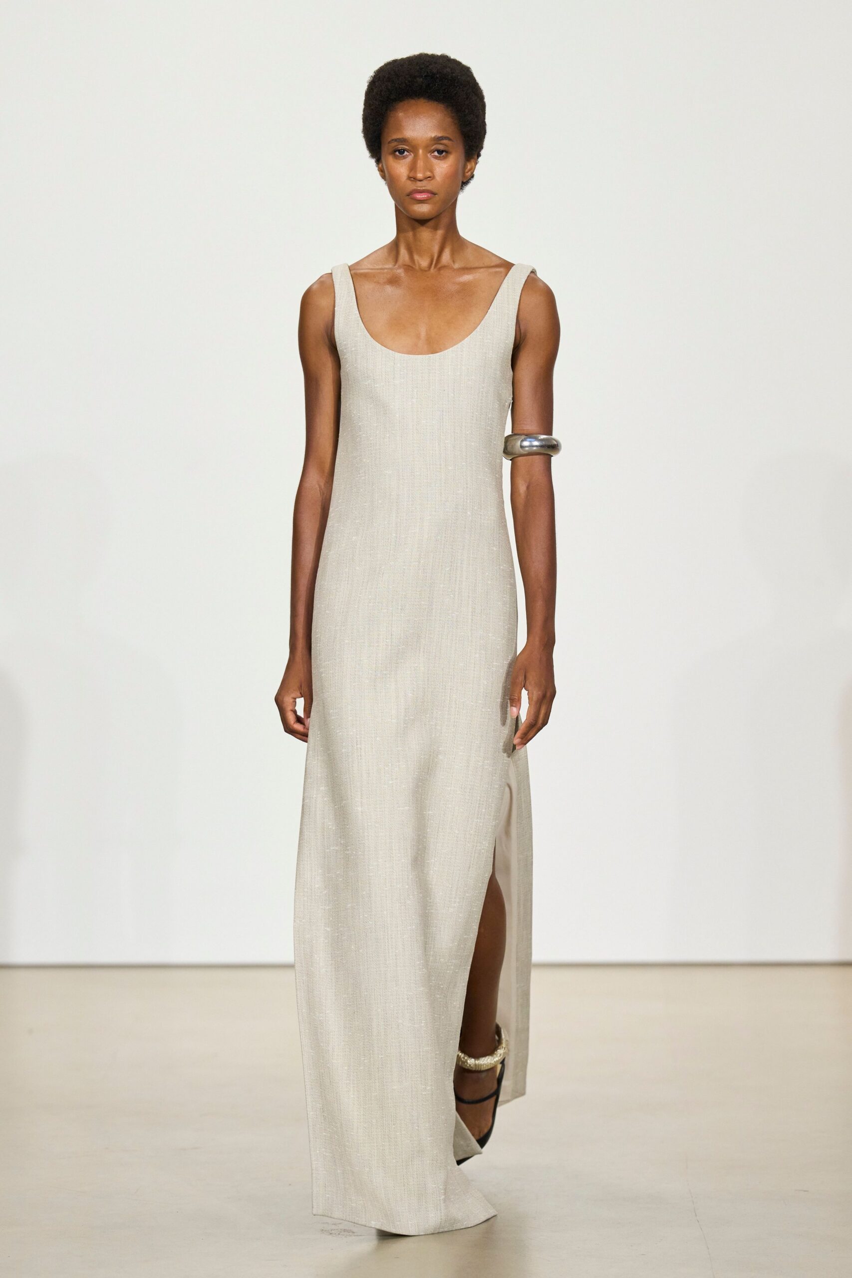 Pale oyster column dress in glinting ribbed knit, deep scoop neck. Oversized silver cuff balanced high on one upper arm. Black ankle-strap heels with a thick braided ring above the foot. Crossing a museum atrium before the floor polish has fully dried.