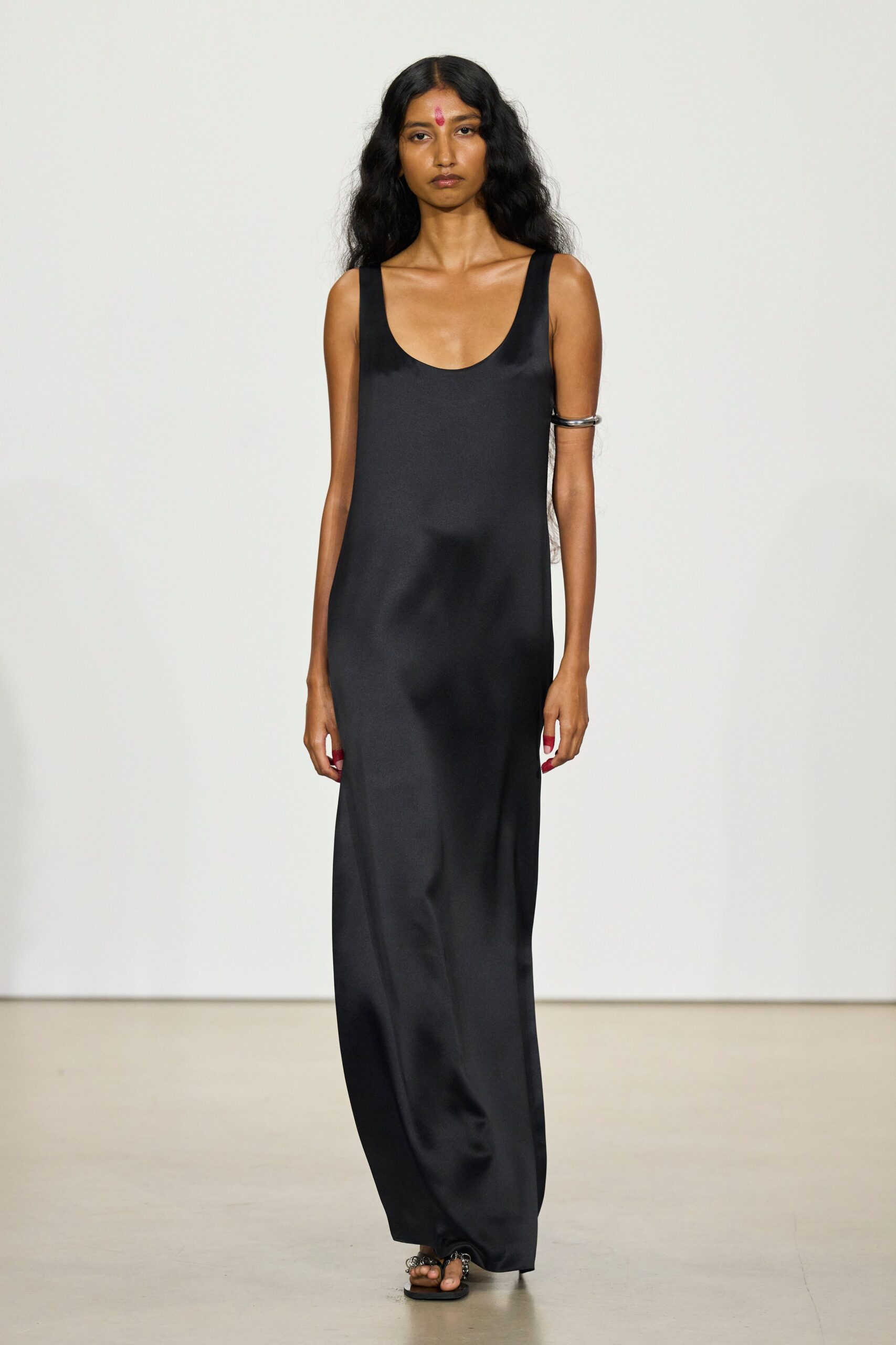 Black satin slip dress cut straight to the floor, wide scooped straps. Thick silver cuff gripping the upper arm. Flat black thong sandals with clustered silver rings at the toes. Temple courtyard at dusk, incense ash still warm on the stone.