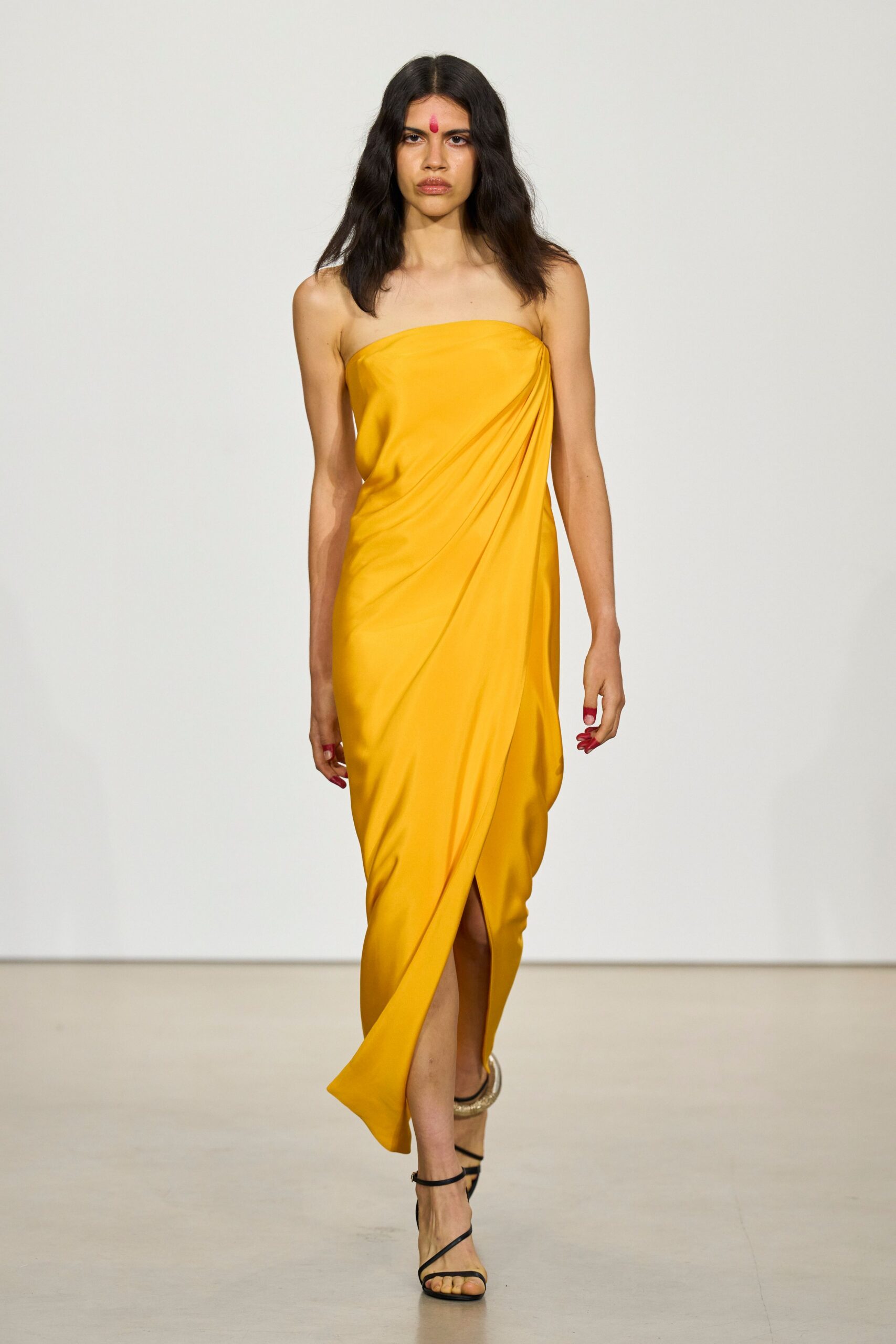 Marigold strapless column dress, diagonally draped with a tulip overlap at the shin. Black barely-there ankle-strap sandals, one ankle stacked with a hammered gold cuff. Temple courtyard wedding, heat still rising from the stone.