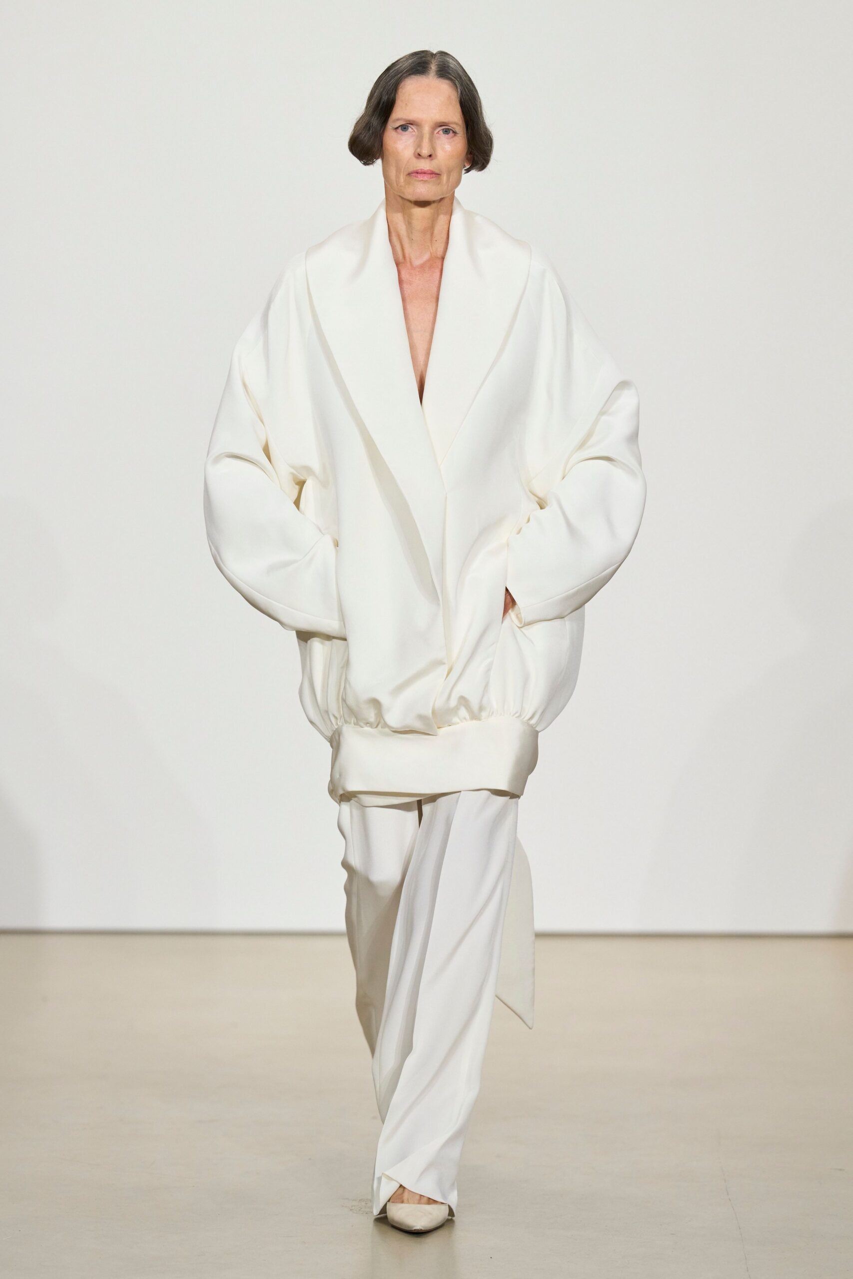 Ivory blouson in heavy crepe, shawl lapels plunging to the sternum. Matching fluid trousers, one front panel crossing over the shoe. Pointed ivory pumps peeking from the hem. For the hushed hour before a courthouse wedding, florist paper still rustling.