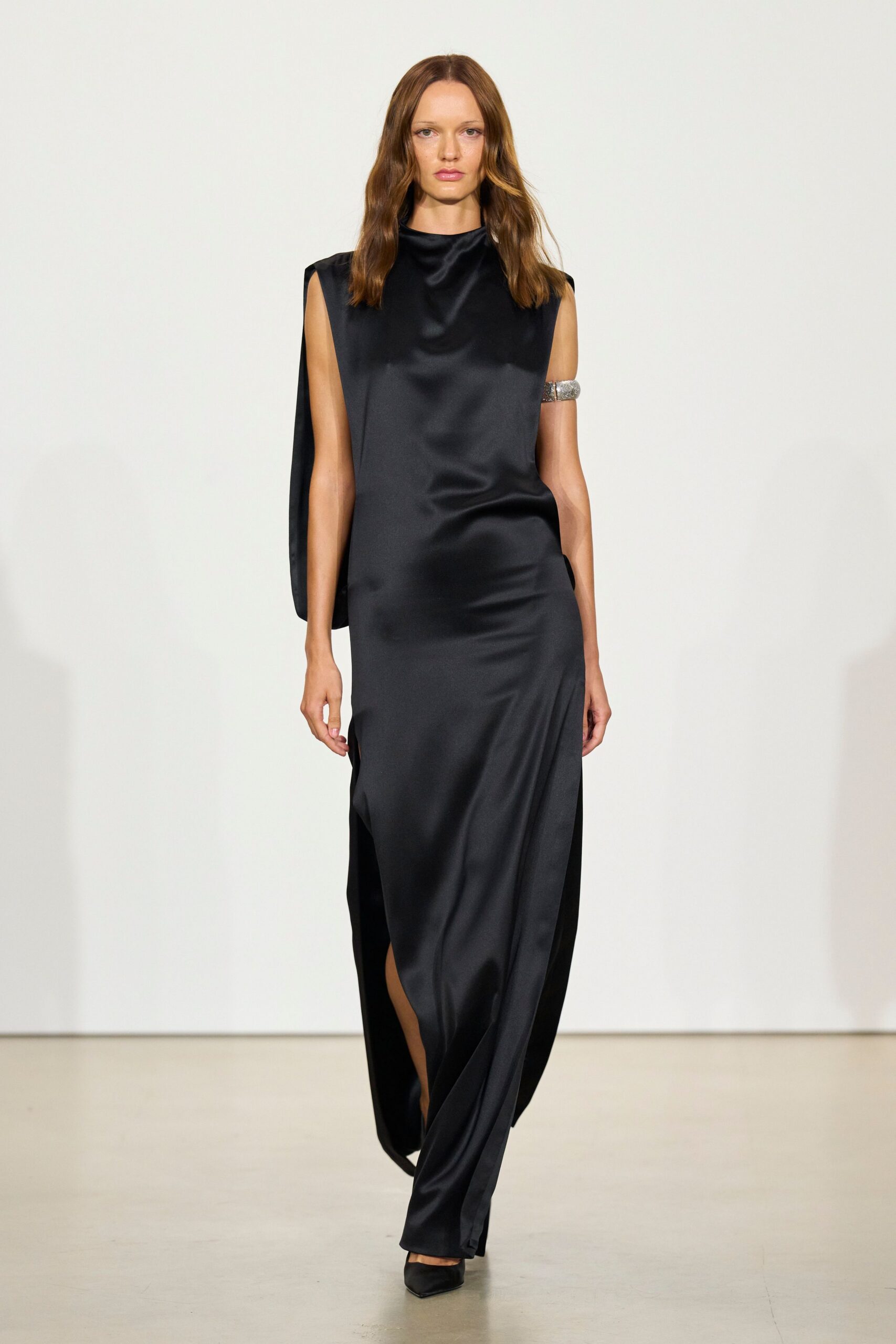 Black satin column gown with padded shoulders and a loose side drape. Silver pavé cuff gripping the upper arm. Black pointed pumps, barely showing under the hem. Hotel corridor after midnight, keycard failing on the third try.