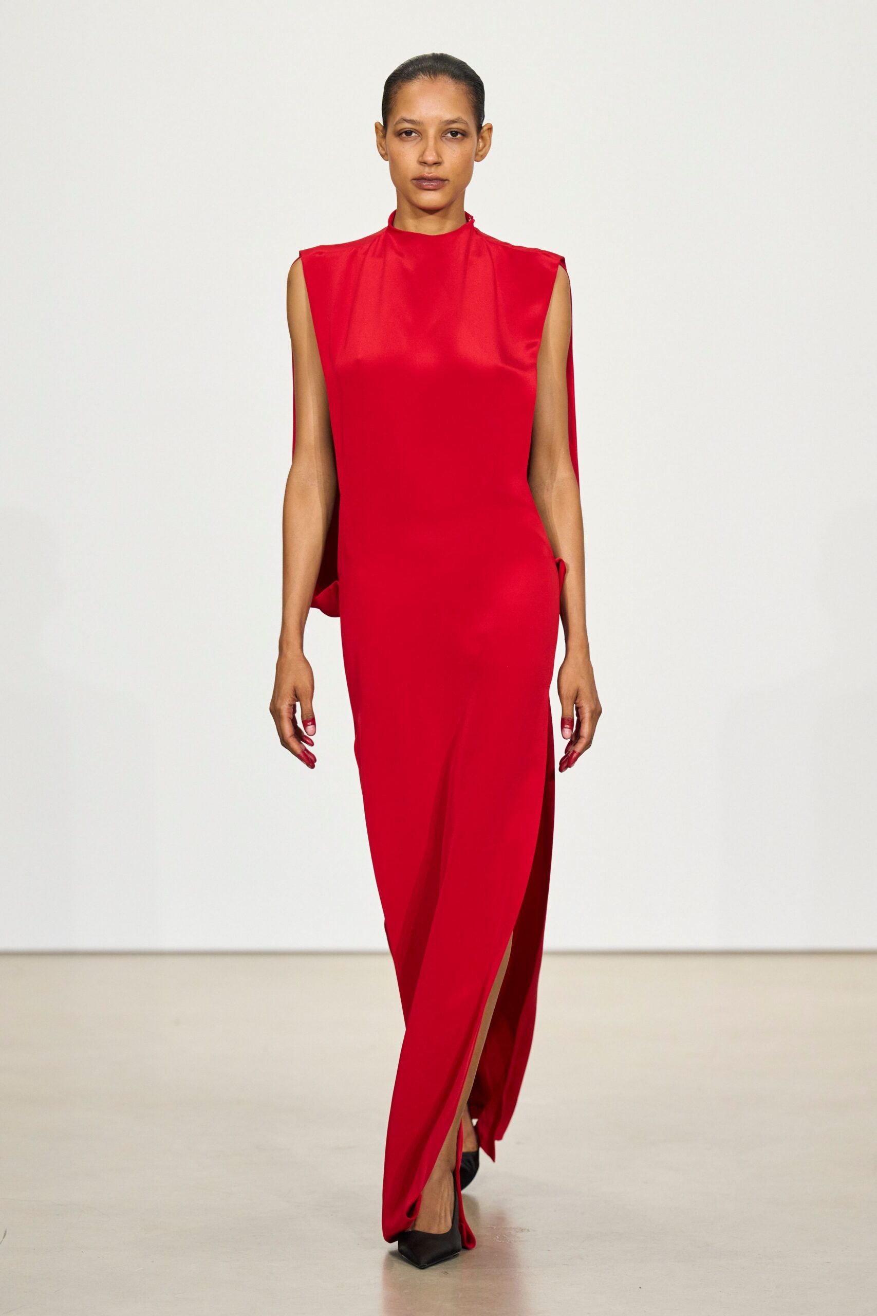 Tomato-red column gown in glossy crepe, with squared cap shoulders and a front slit. Black pointed pumps, nearly hidden under the dragging hem. Crossing a hotel lobby before sunrise with yesterday's lipstick still on.