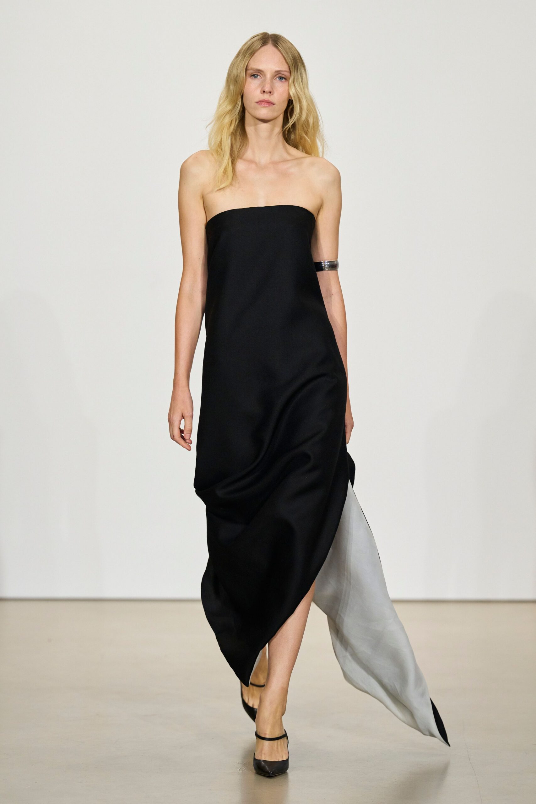 Black strapless column dress in heavy satin, one side releasing a white organza train. Wide silver cuff clasping the upper arm. Black pumps with a fine ankle strap. Crossing a museum atrium after the rain stopped, carrying silence with you.
