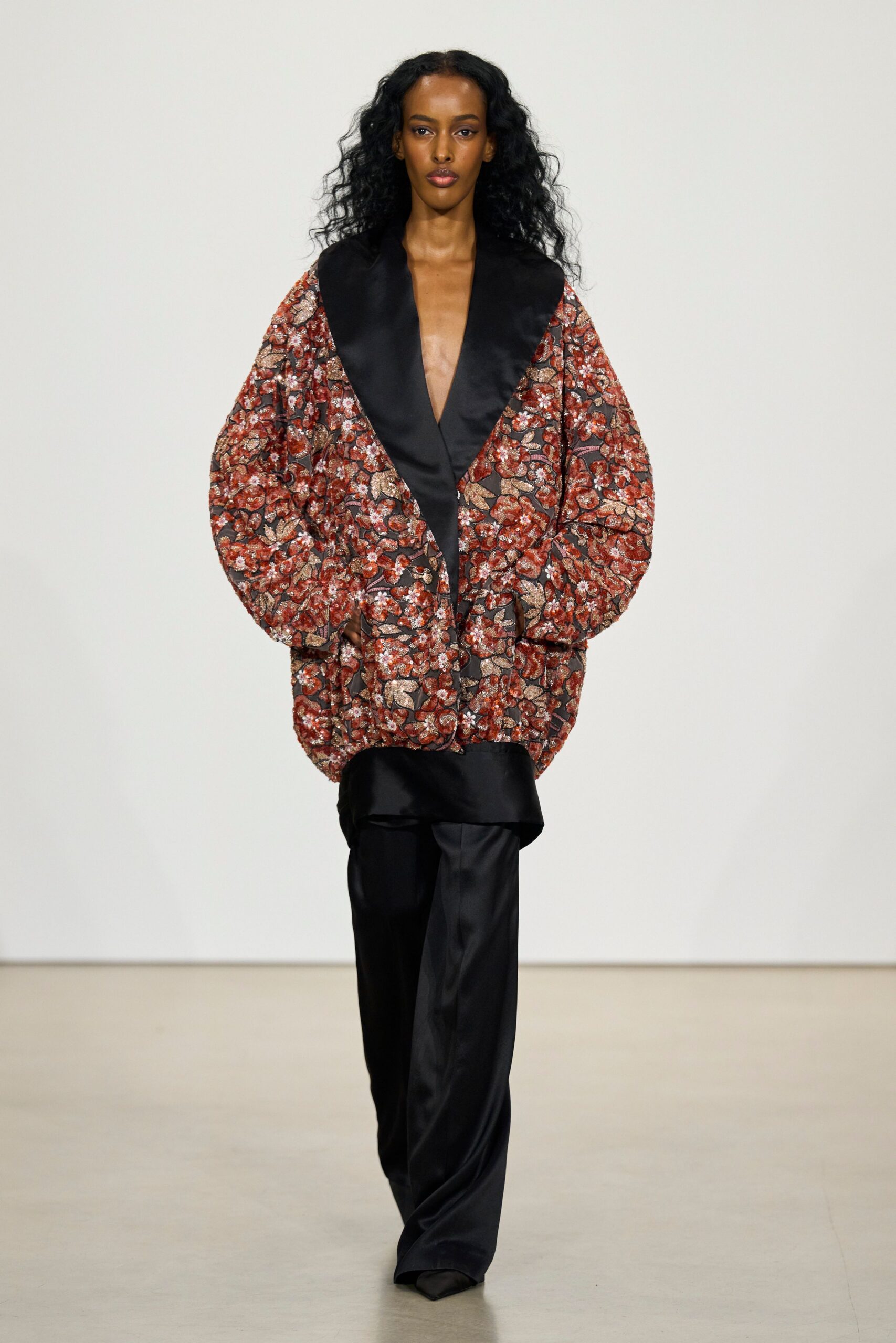 Oversized brocade jacket in rust floral weave with a deep black satin shawl collar. Black satin tunic, hem peeking below the jacket in a soft curve. Fluid black satin trousers falling long over pointed toes. Hotel corridor at dawn, still carrying last night's perfume.
