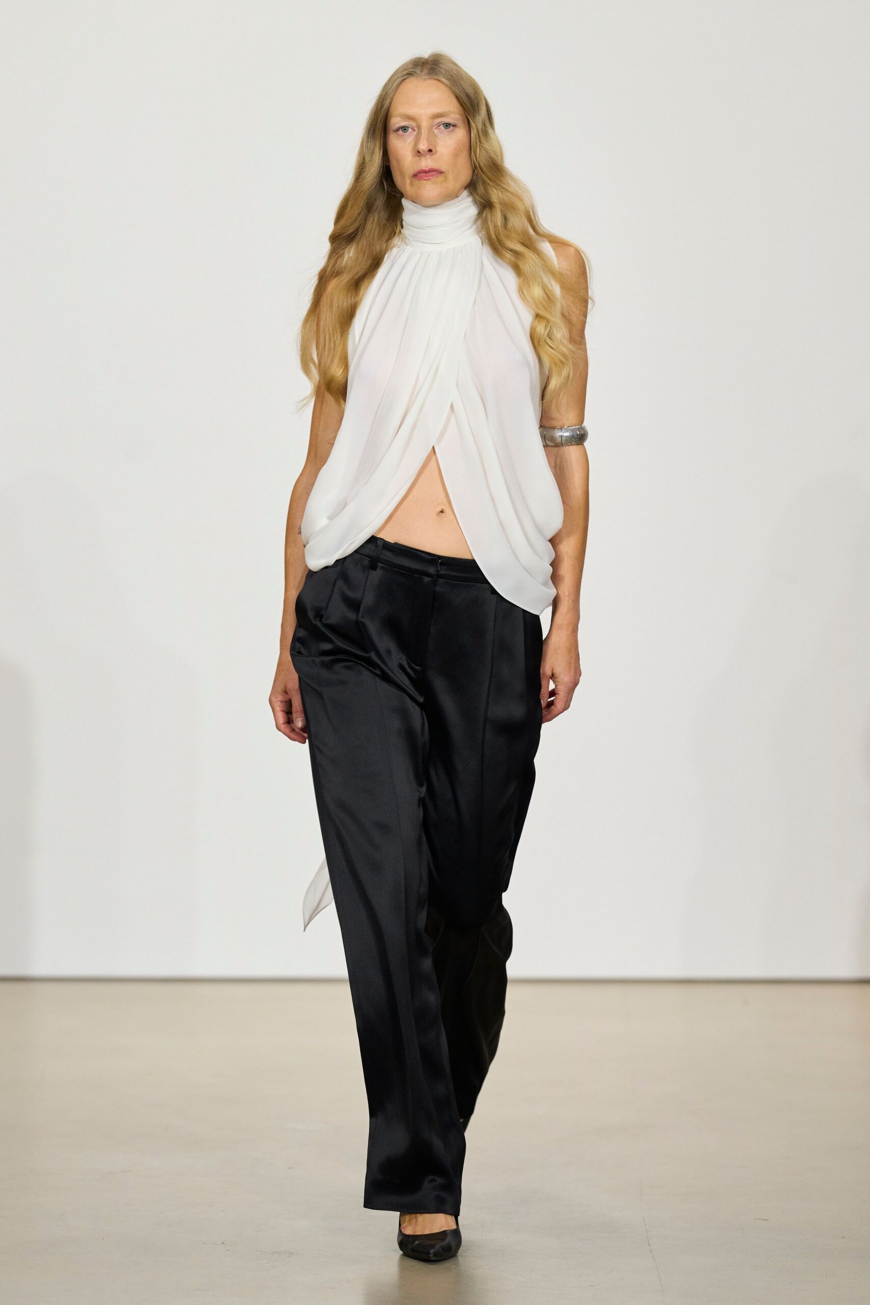 Ivory georgette halter with a ruched turtleneck and split draped front. Wide silver cuff hugging the upper arm. Black satin trousers, sharply creased with a low tab fastening. Black leather heels with squared-off toes. Hotel corridor at dawn, carrying your shoes back from someone else's room.