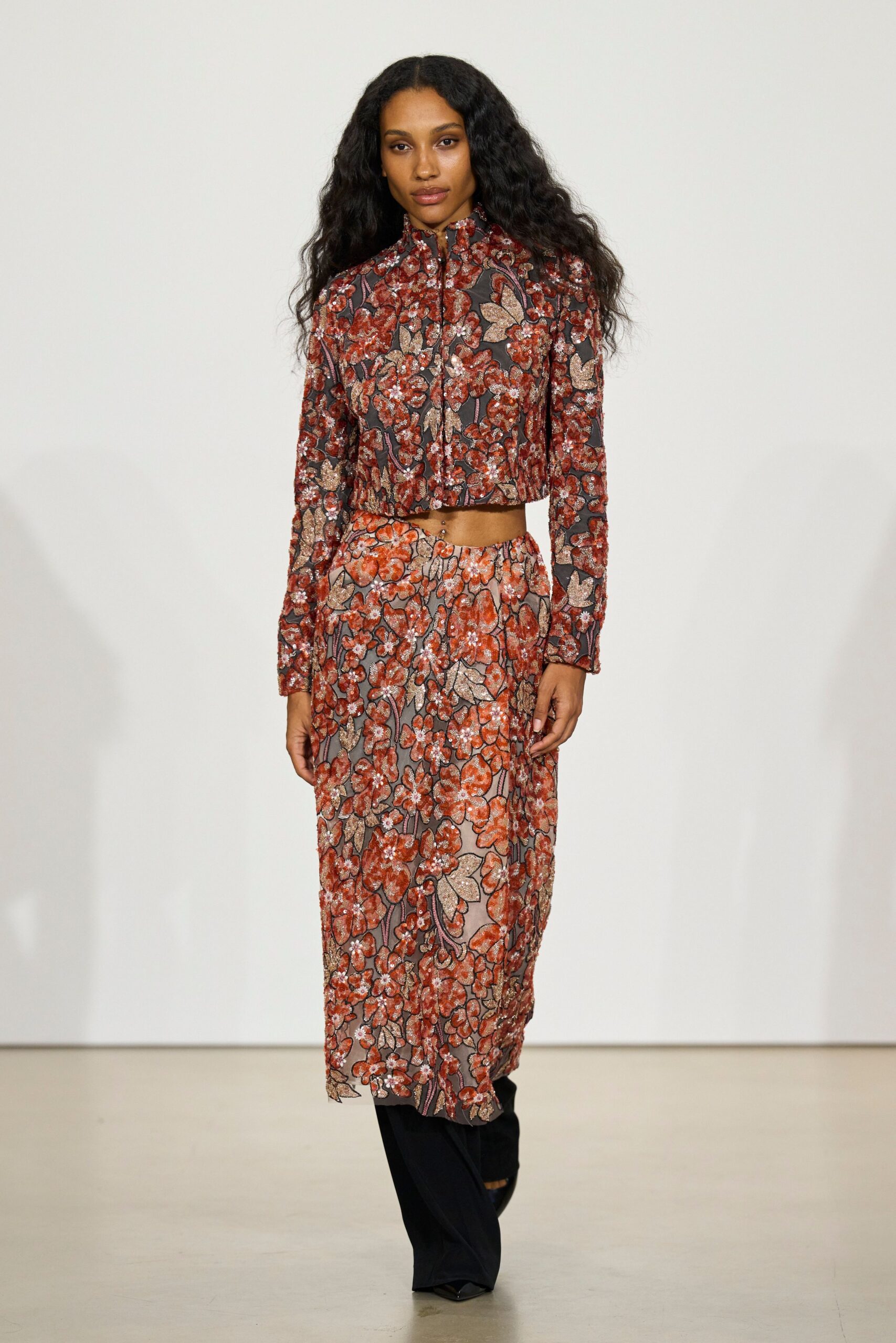Cropped zip-front jacket in rust floral sequins, collar standing close. Matching column skirt, slit opening off-center from the hem. Black trousers breaking wide beneath the sequined skirt. Walking into a wedding reception after the church part ran long.
