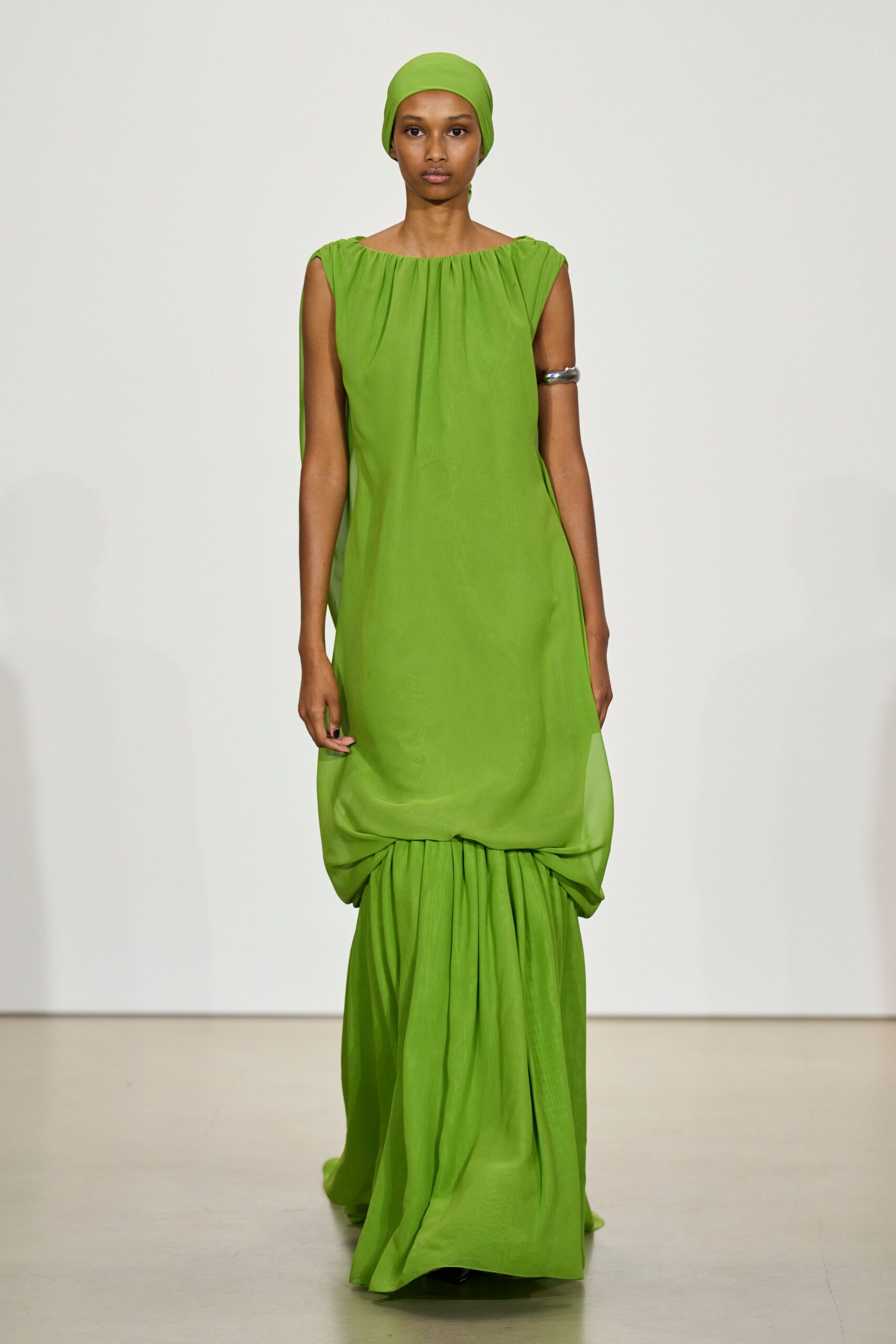 Lime jersey headscarf pulled smooth and close over the crown. Lime column dress in fluid crepe, neckline softly gathered and skirt caught into a low bubble. Wide silver cuff hugging the upper arm. Heat shimmering off museum stone at noon.