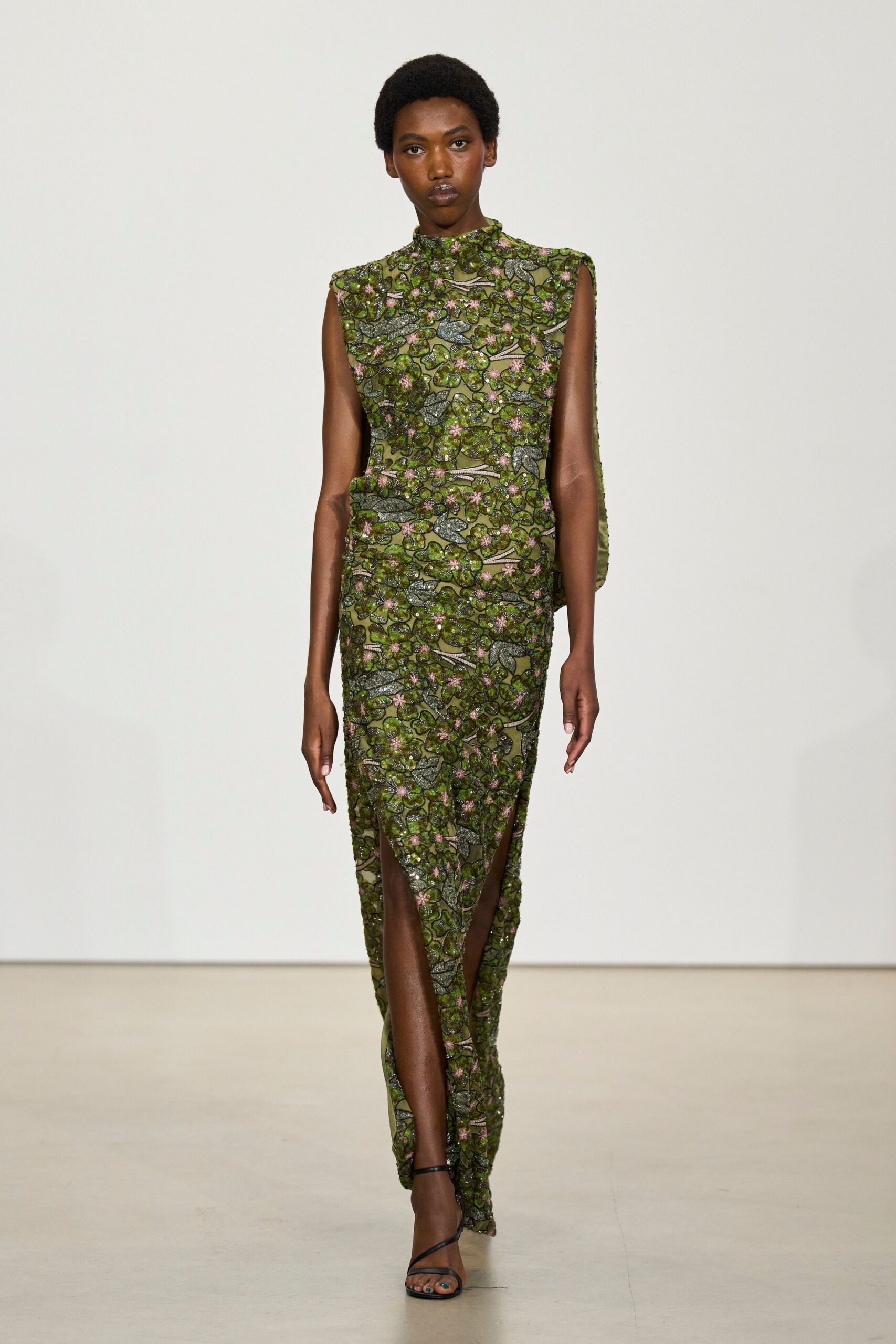 Olive mesh column dress densely beaded with vine leaves and pink blossoms, split high at the shins. Black barely-there sandals with thin wraparound straps. Matching beaded pouch tucked under one arm. A conservatory wedding after rain, shoes clicking on wet stone.