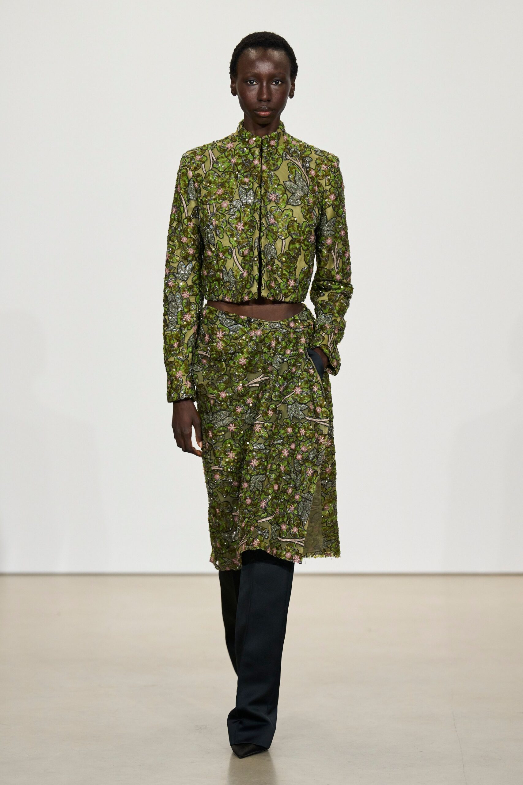 Olive mesh jacket densely beaded with silver leaves and pink blossom clusters. Matching beaded skirt, split open at one thigh with a softly frayed hem. Black trousers flaring past the skirt into sharp pointed shoes. Walking out of a conservatory fundraiser with damp petals on the path.