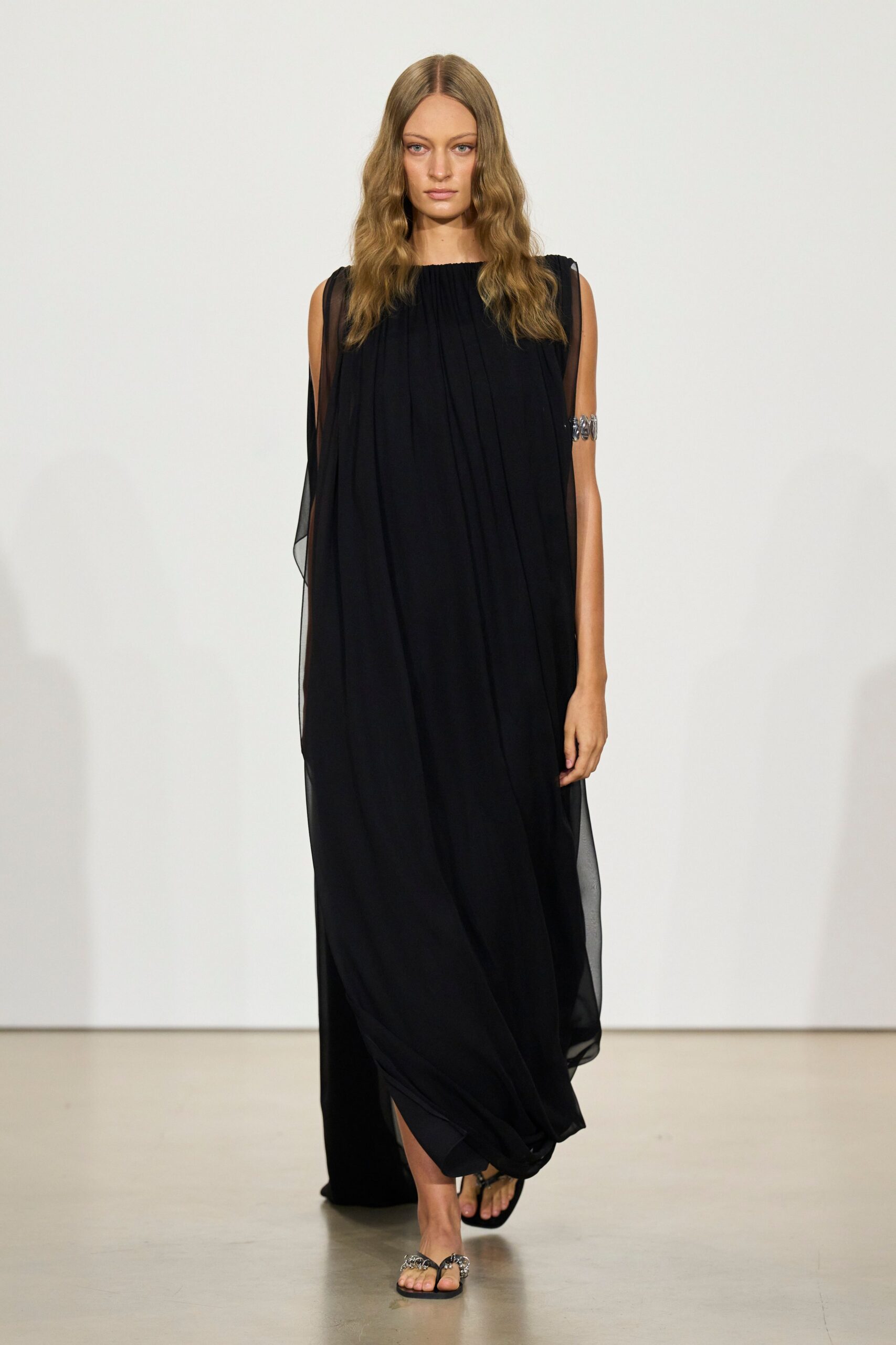 Black chiffon column dress, softly gathered at the neck with sheer side drapes. Silver oval-link cuff gripping the bare upper arm. Flat black thong sandals with jeweled loops over the toes. Walking through a whitewashed villa after midnight, floor still warm underfoot.