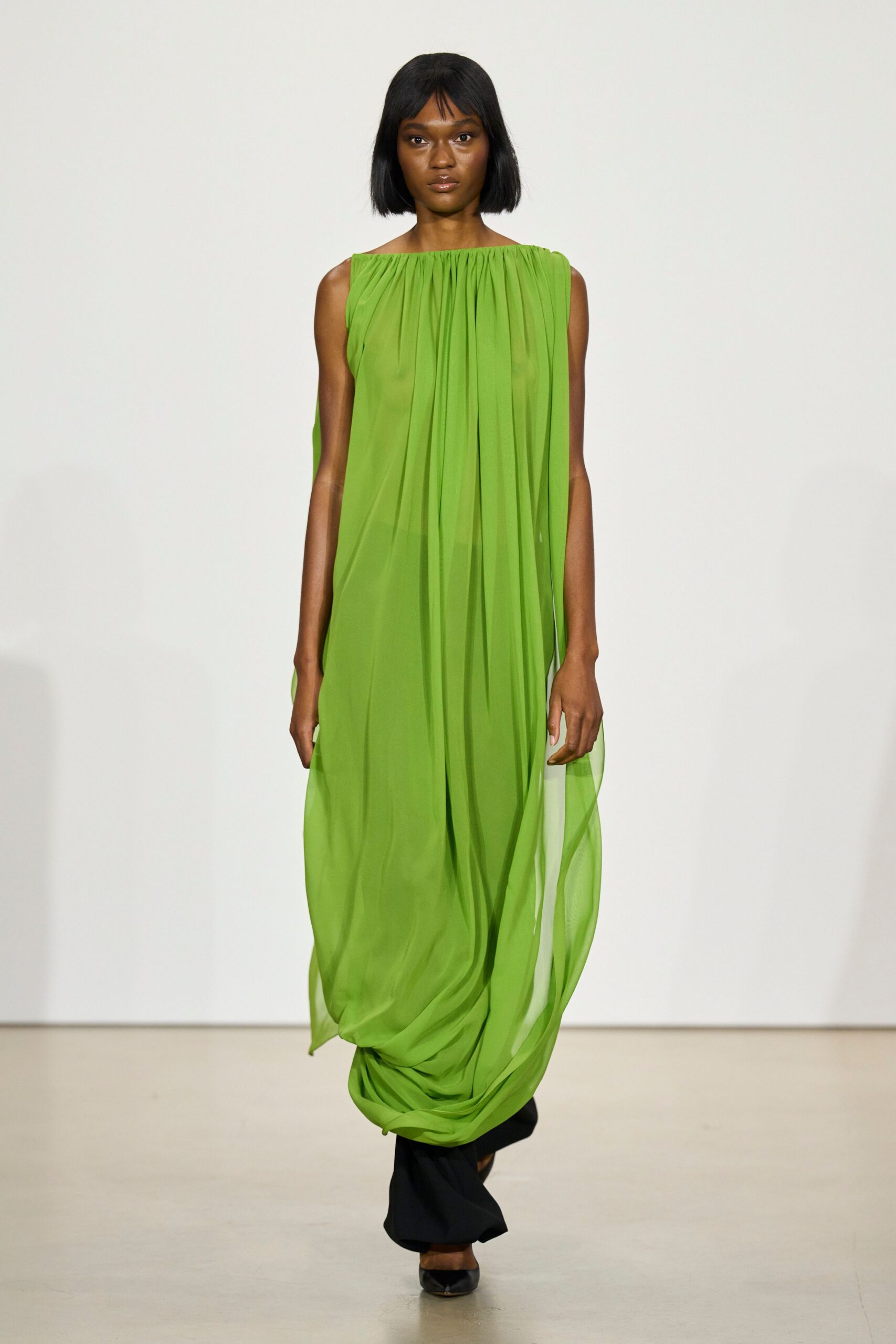 Acid-green chiffon column, densely gathered at the boatneck and puddling low. Black flared trousers peeking from the dress slit. Pointed black pumps with a narrow glossy toe. Crossing a museum atrium with wet flowers wrapped in paper.