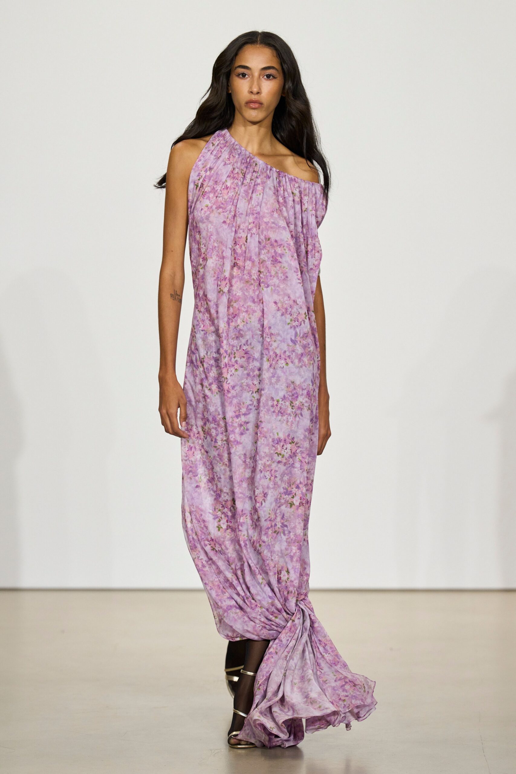 Lavender floral chiffon gown, one-shoulder neckline gathered into loose vertical drape. Metallic barely-there sandals with slim straps across the toes. Crossing a hotel terrace after the wedding, hem already sweeping fallen petals.