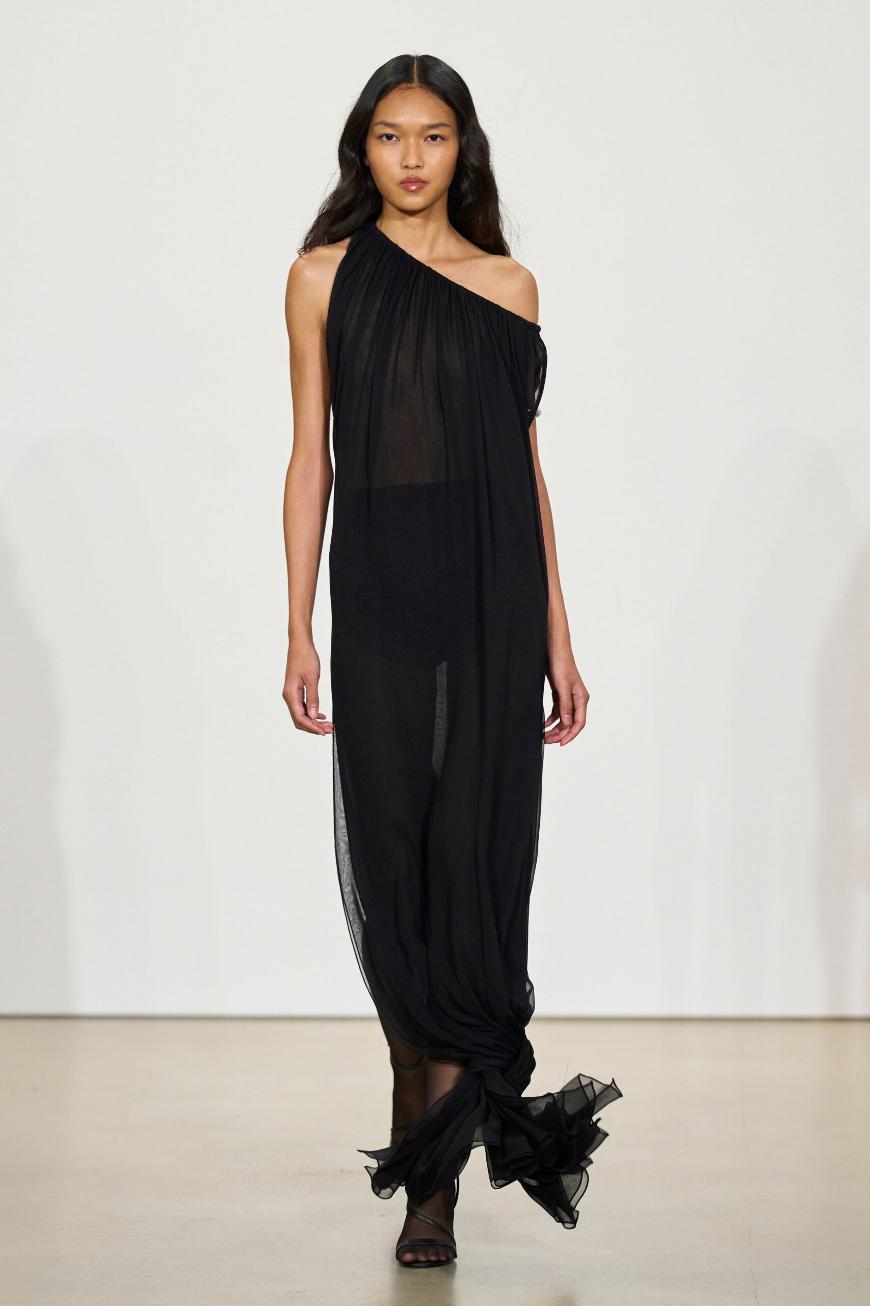 Black chiffon gown draped one-shoulder, sheer through the torso and knotted at the hem. Black barely-there sandals under a smoky sheer skirt. Whatever trails behind you leaving a hotel wedding after midnight.
