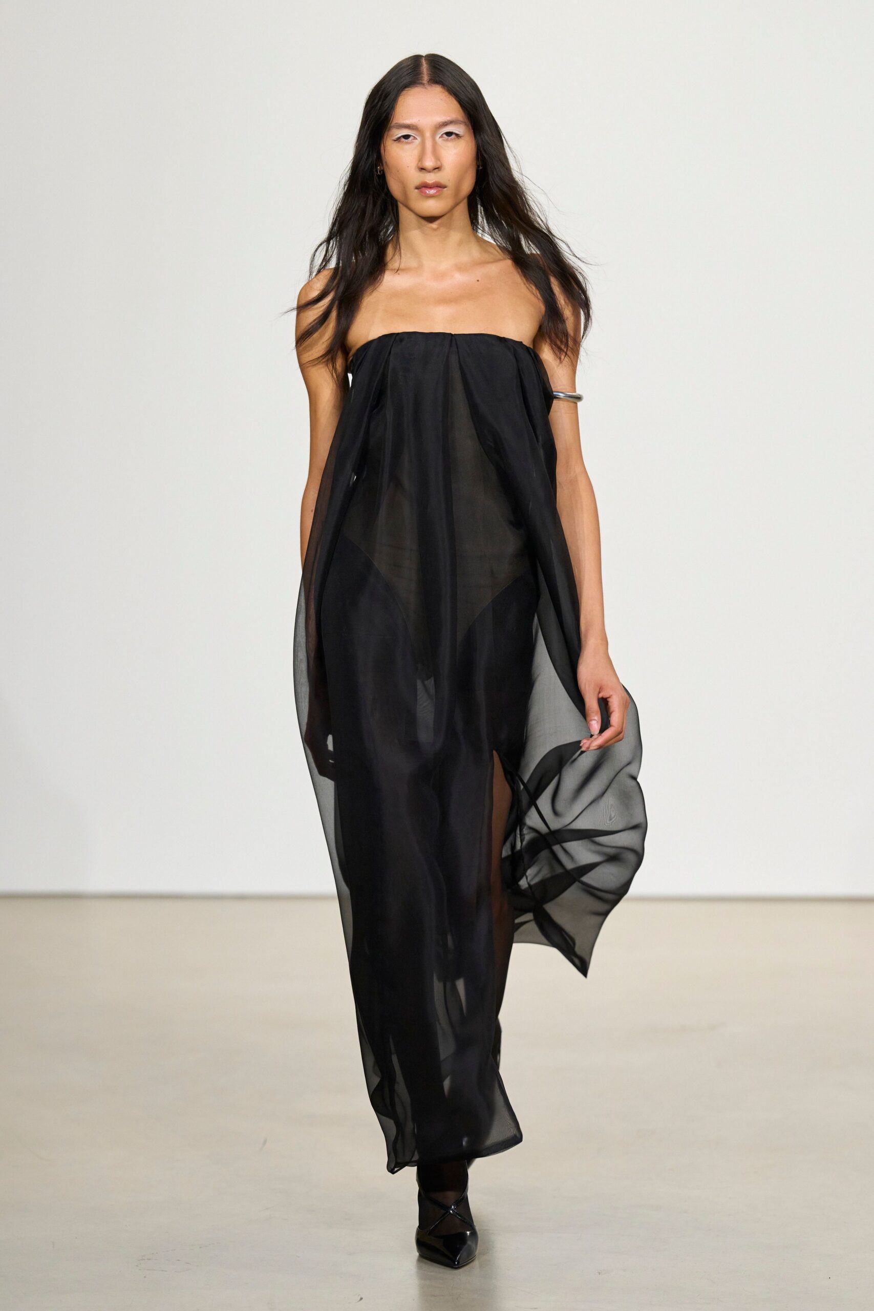 Black strapless chiffon column, softly gathered at the top edge. Thick silver cuff riding high on one bare upper arm. Black pointed flats with crossed straps and a glossy toe. For opening the terrace door after midnight rain.