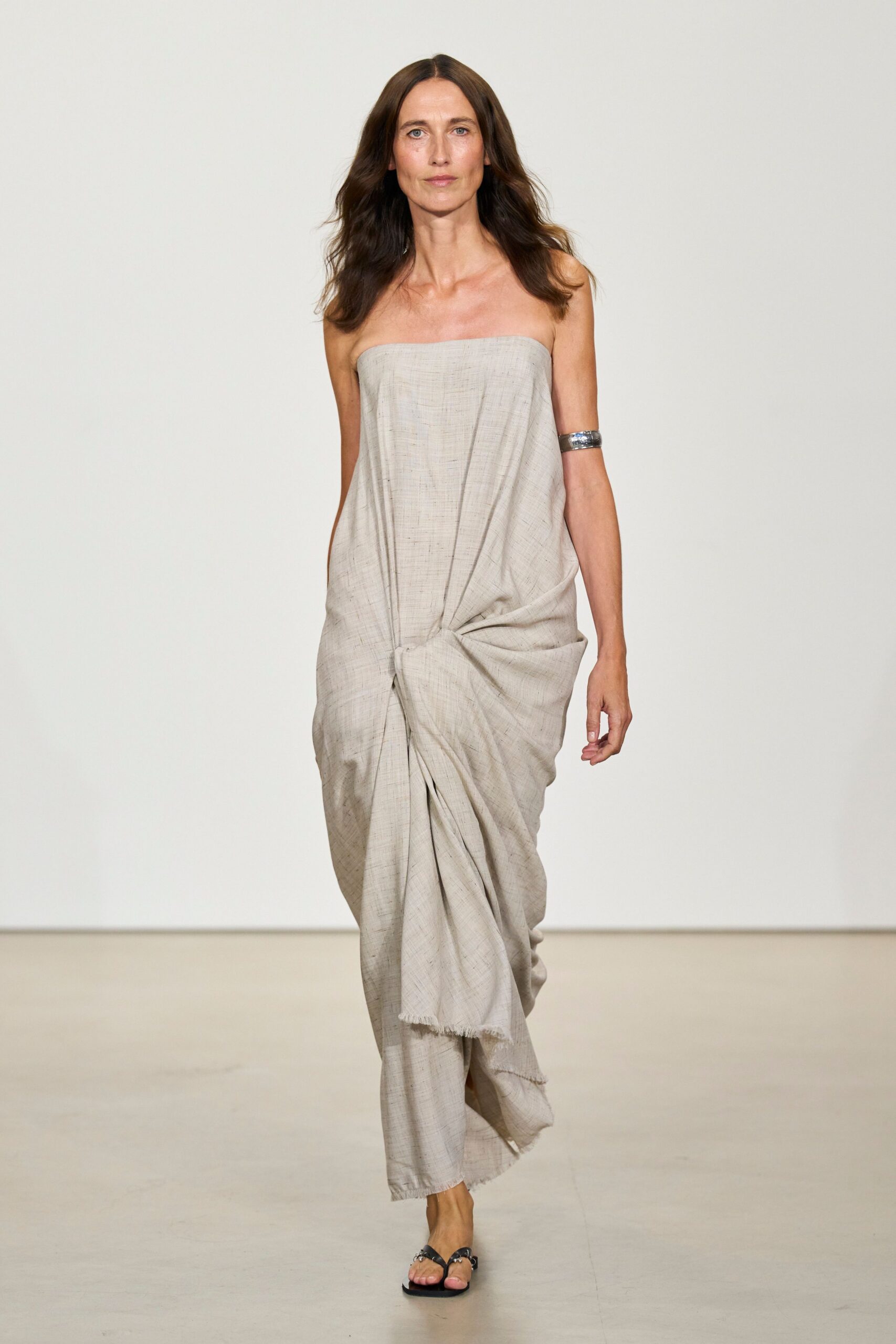 Stone slub-weave strapless dress, twisted into a low knot at the hips. Wide hammered silver cuff on the upper arm. Black thong sandals with tiny rings at the straps. Walking barefoot after dinner until the boardwalk planks turn cold.