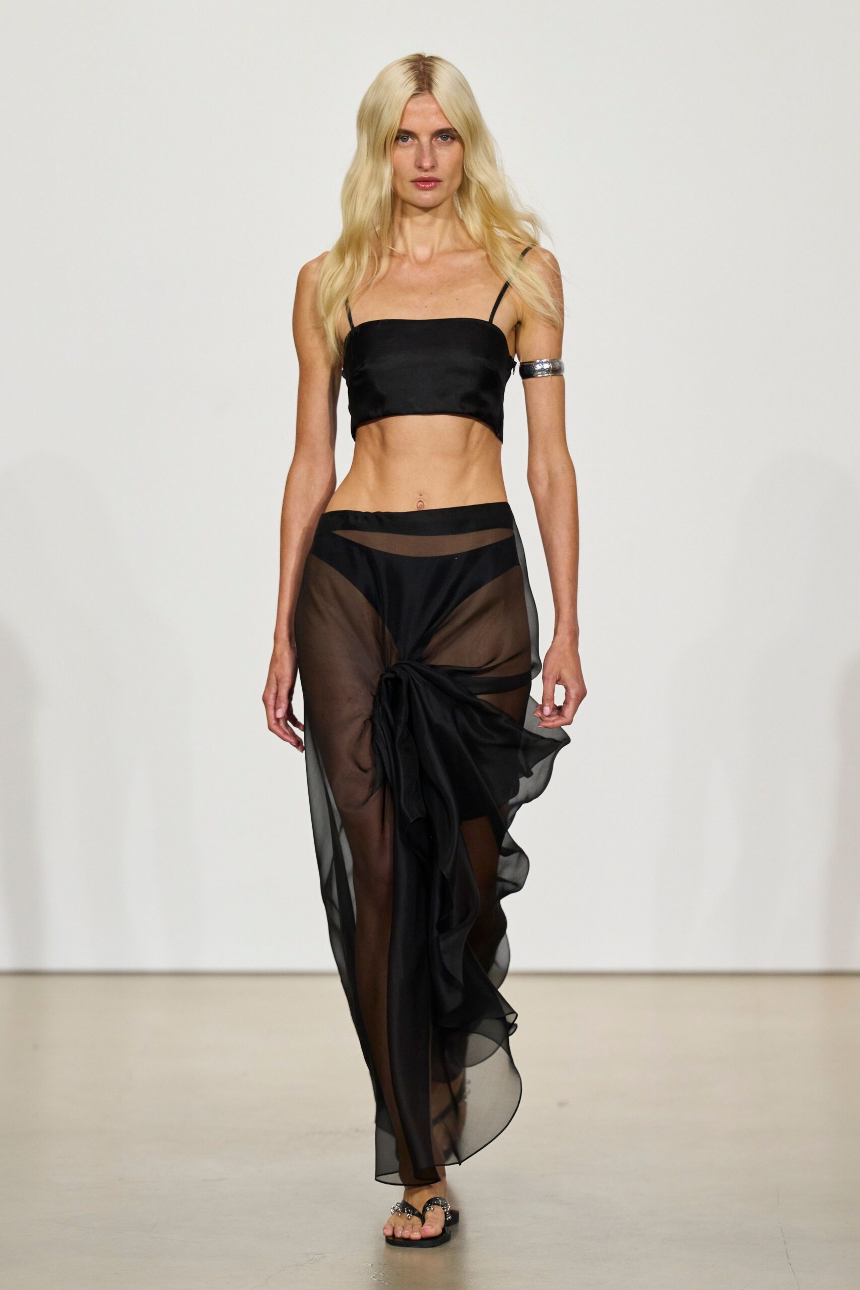 Black satin bandeau crop top with thin spaghetti straps. Wide silver cuff, worn high on one bare upper arm. Sheer black chiffon sarong skirt knotted low, brief peeking through. Flat black thong sandals with clustered silver charms at the toes. Walking back from the hotel pool after midnight, still salty at the collarbone.