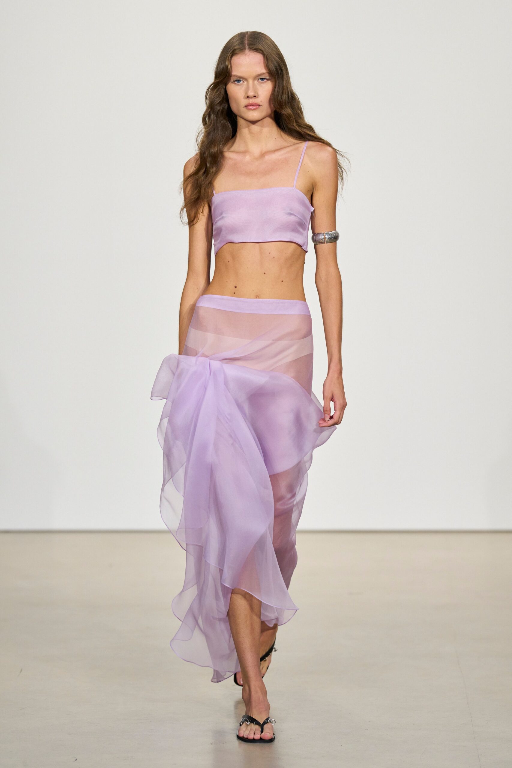 Lilac satin crop top with a shallow curved hem. Wide silver cuff, dented like hammered metal. Sheer lilac organza wrap skirt knotted low in a swollen side bow. Black thong sandals with tiny silver rings at the strap. Walking the length of a pool terrace before anyone gets in.