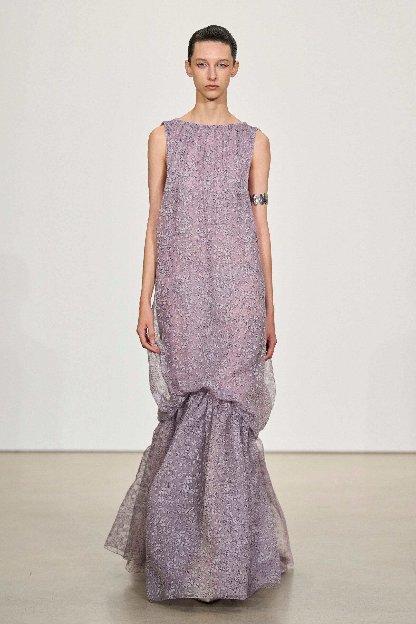 Mauve sheer gown in tiny floral lace, dropped into a bubbled train at the knees. Wide silver cuff of oval links gripping the upper arm. Crossing a museum atrium after the florist has swept the floor.