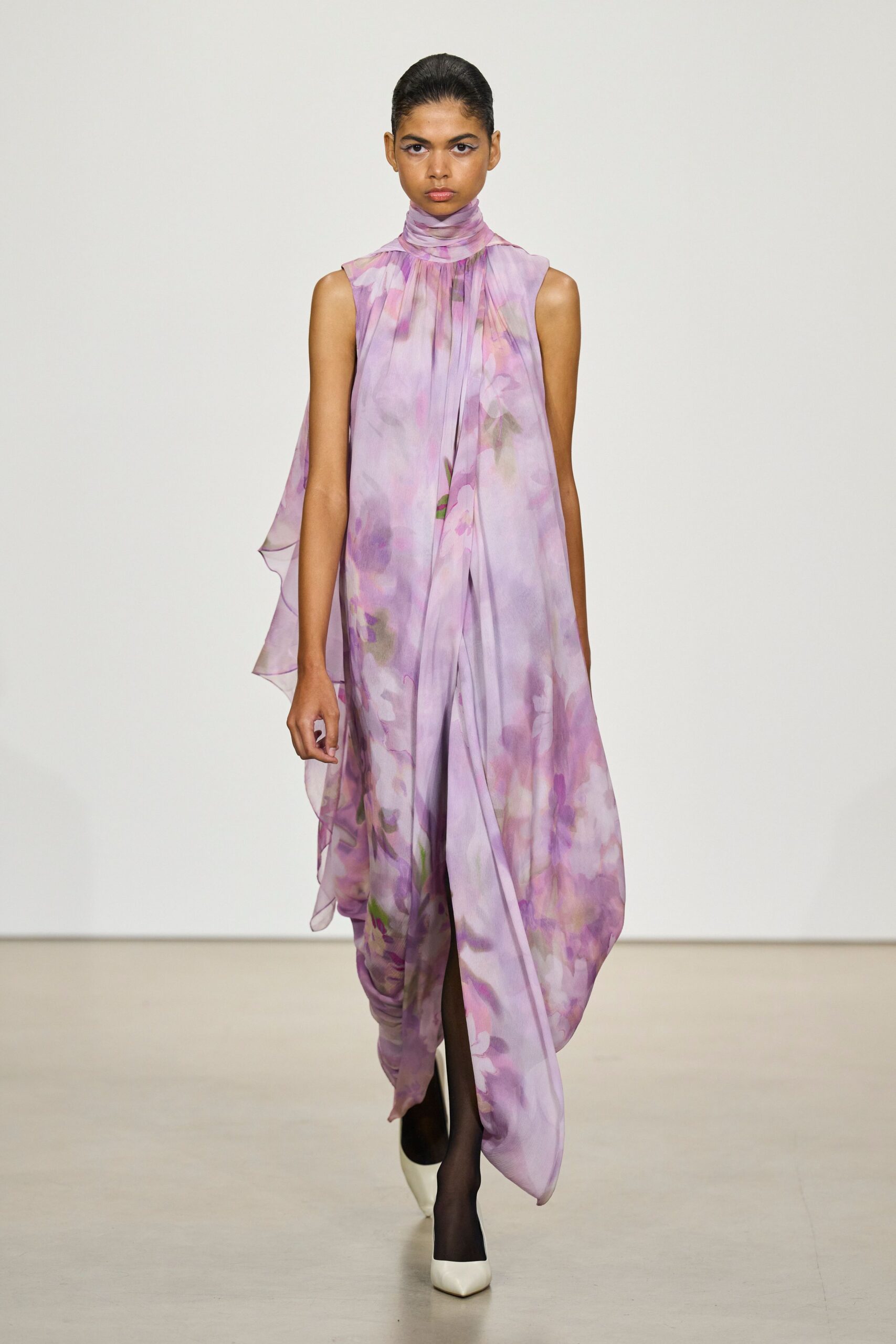 Lavender watercolor chiffon dress, high wrapped neck and soft gathered plunge. Sheer black tights. White pointed pumps under the draped hem. Crossing a botanic conservatory just after the glass has fogged over.