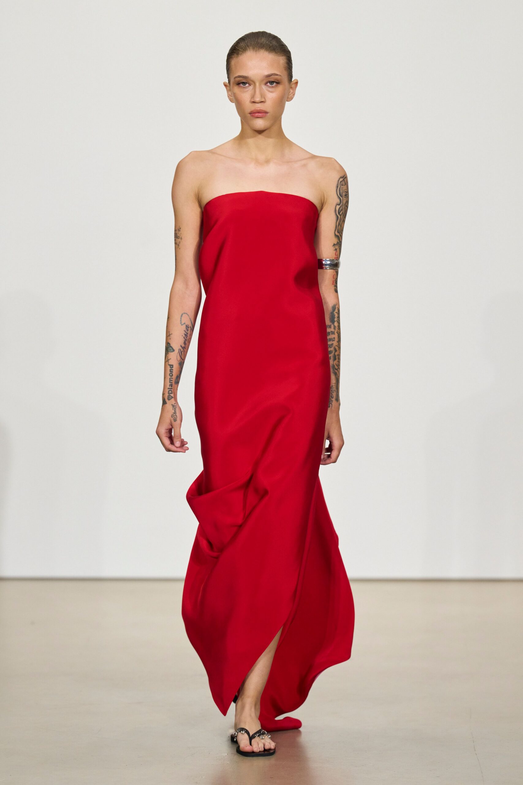 Strapless scarlet column dress in fluid satin, one heavy fold buckling the skirt. Wide silver cuff gripping the upper arm. Black thong sandals with jewel-cluster straps. Hotel wedding after midnight, carrying your shoes back across the marble lobby.