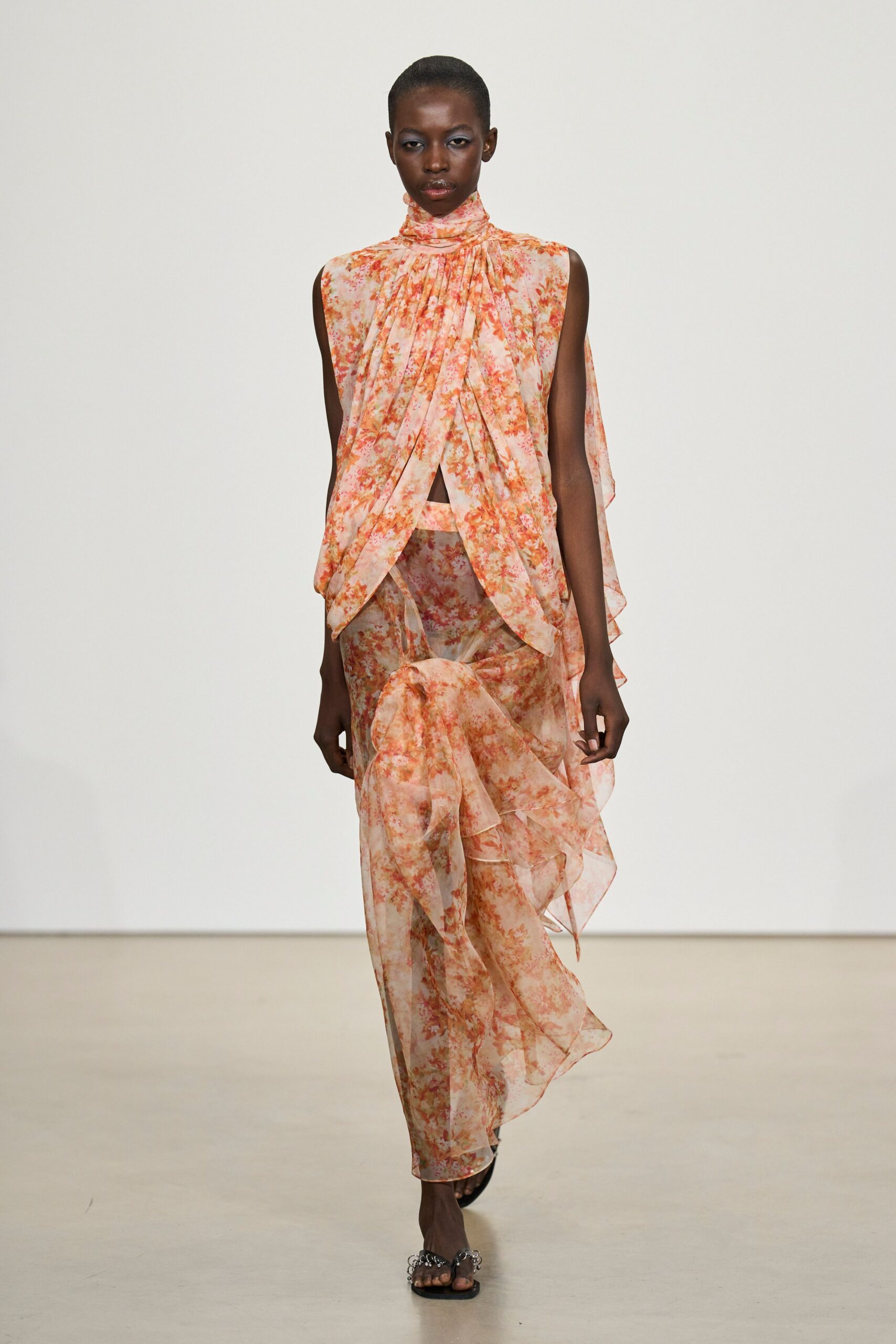 Blush chiffon blouse in a washed coral floral, high neck softly ruched. Matching sheer chiffon skirt, draped into a knotted cascade at one knee. Black thong sandals with clustered silver rings across the toes. Walking out to a hotel terrace just after the garden wedding ends.