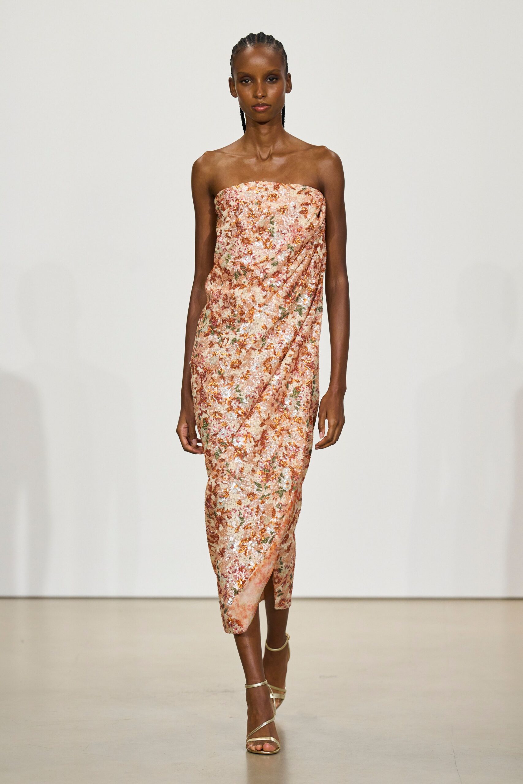 Strapless column dress in blush sequin floral, draped diagonally into a wrapped hem. Bare gold ankle-strap sandals. Champagne on a terrace after the wedding guests have gone downstairs.