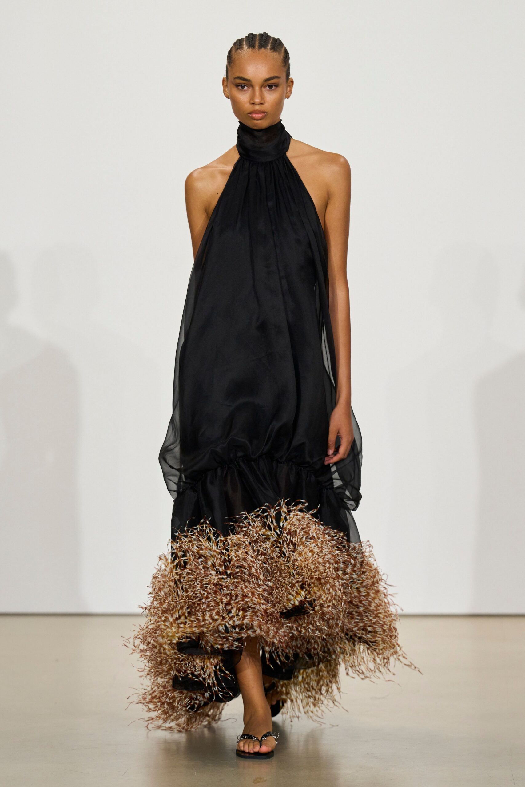 Black halter dress with a scarf neck, sheer side panels and straw-fringe hem. Flat black thong sandals with a small silver ring at the toe. Crossing a hotel courtyard after midnight, hem full of dry ornamental grass.