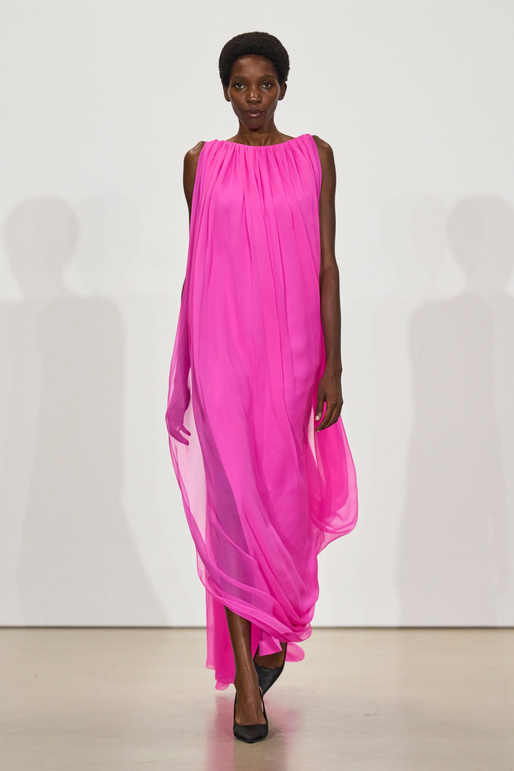 Hot-pink chiffon gown, finely pleated from the collar into loose vertical falls. Black pointed pumps under the pooling hem. Crossing a hotel atrium while the florist is still sweeping petals.