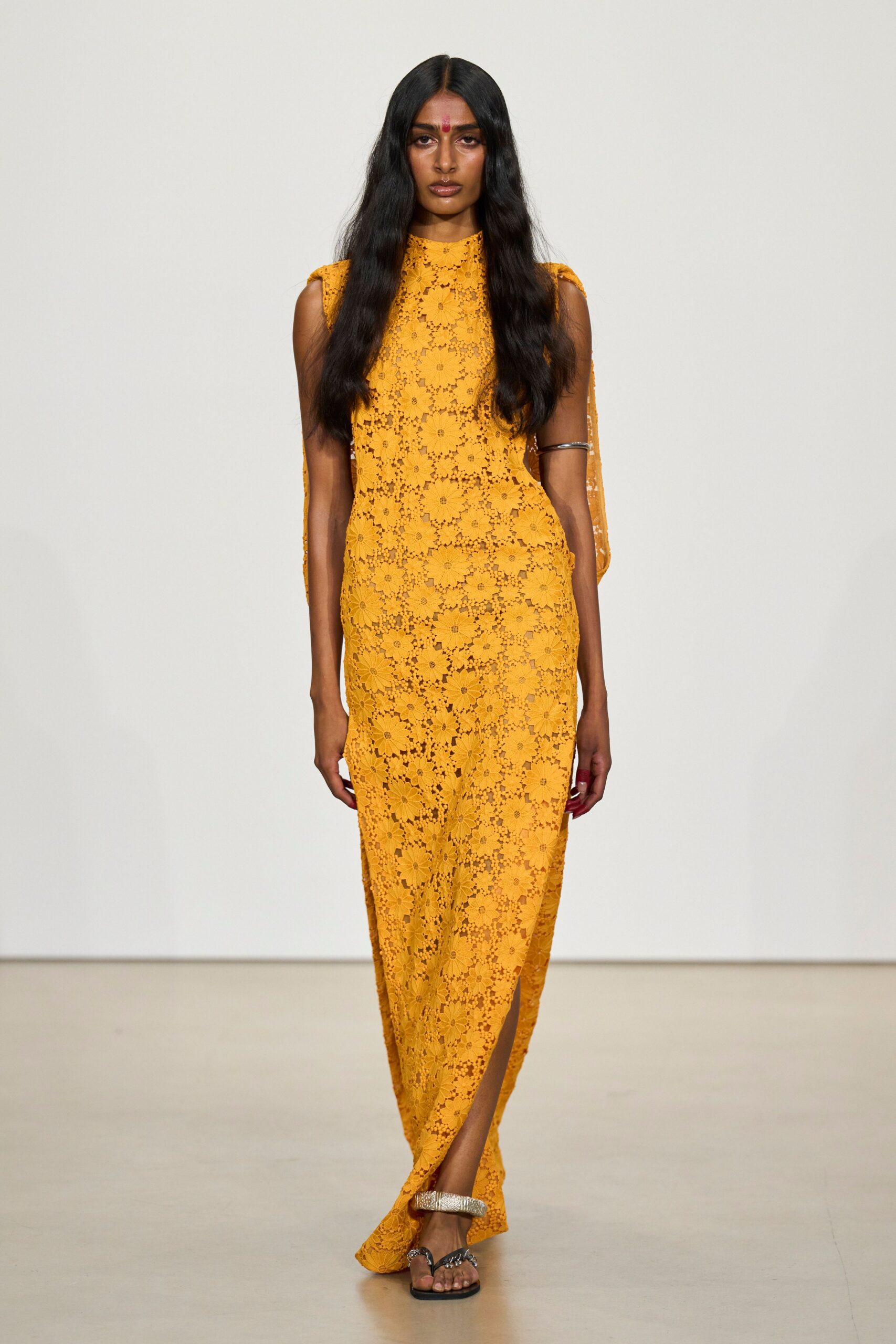 Marigold floral guipure column dress, shoulder panels falling into cape-like strips. Plain silver arm cuff, riding high on the upper arm. Flat black thong sandals with a hammered silver toe strap. Temple courtyard at noon, heat caught in the stone.