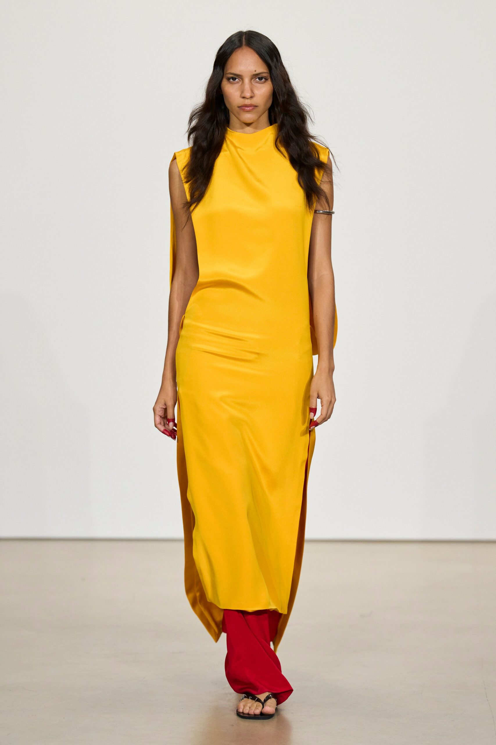 Marigold column dress in liquid satin, slit sides revealing a long back panel. Scarlet trousers pooling softly beneath the hem. Single silver cuff hugging the upper arm. Black thong sandals with a square flat sole. Crossing a museum atrium with flowers still wrapped in paper.