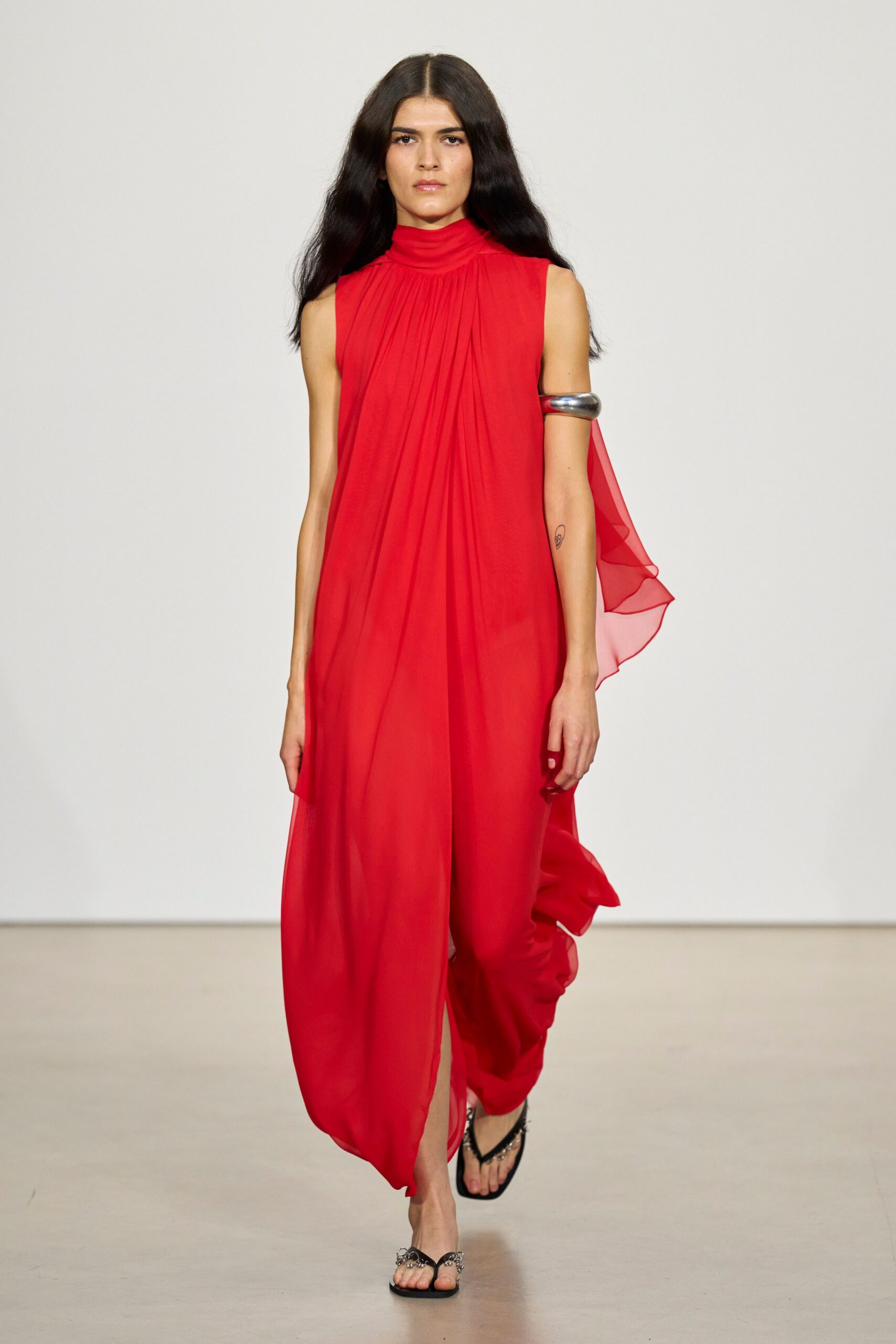 Poppy chiffon column dress, high rolled neck and radiating gathers. Sheer back panel floating loose from one armhole. Oversized silver cuff gripped high on the upper arm. Black thong sandals with clustered metal at the toe. Crossing a hotel atrium while the revolving door keeps catching warm wind.