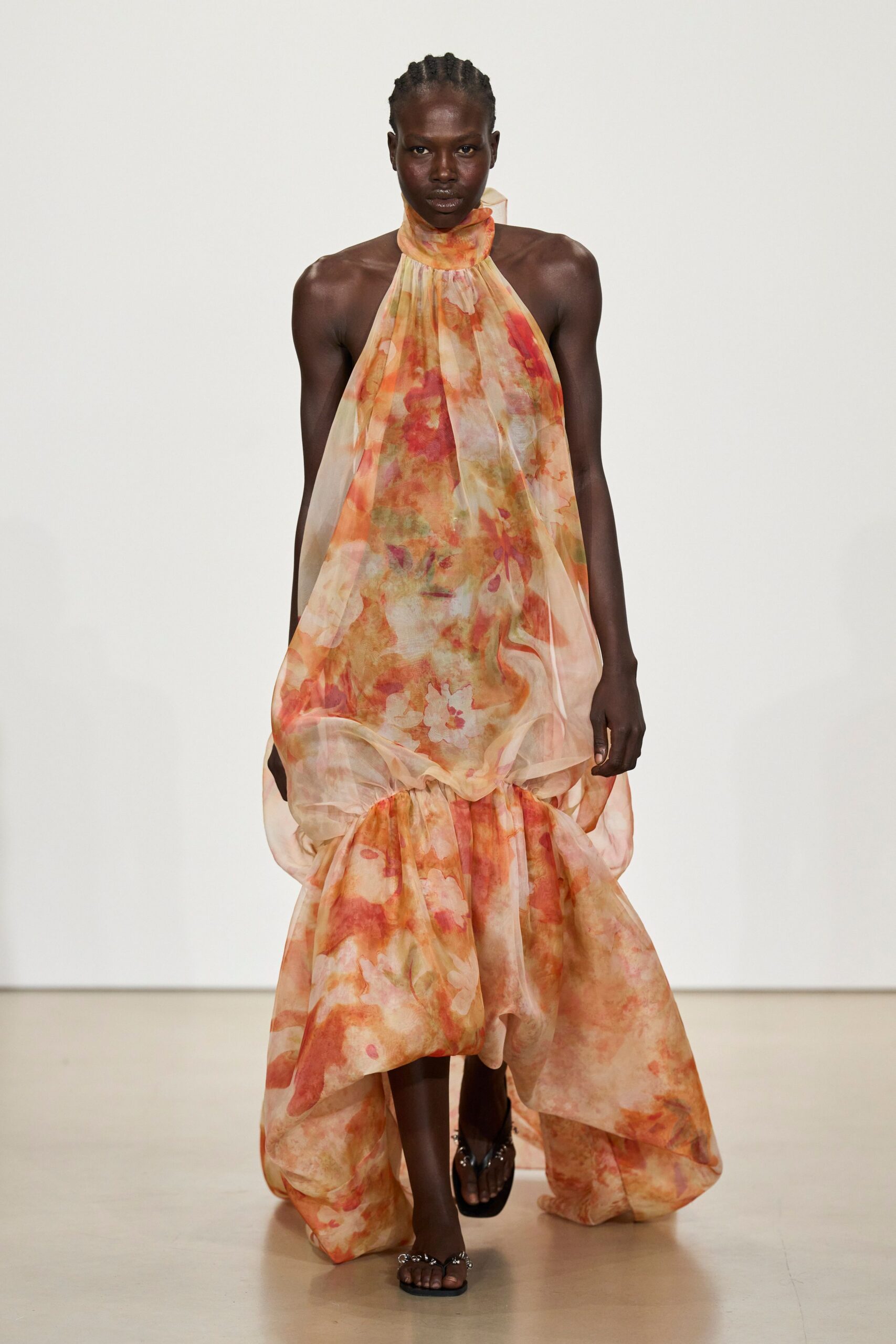 Sheer peach organza halter gown, watercolor florals swelling into a dropped bubble hem. Black thong sandals with clustered silver hardware at the straps. Crossing a hotel courtyard after the wedding, carrying the last heat of sunset.