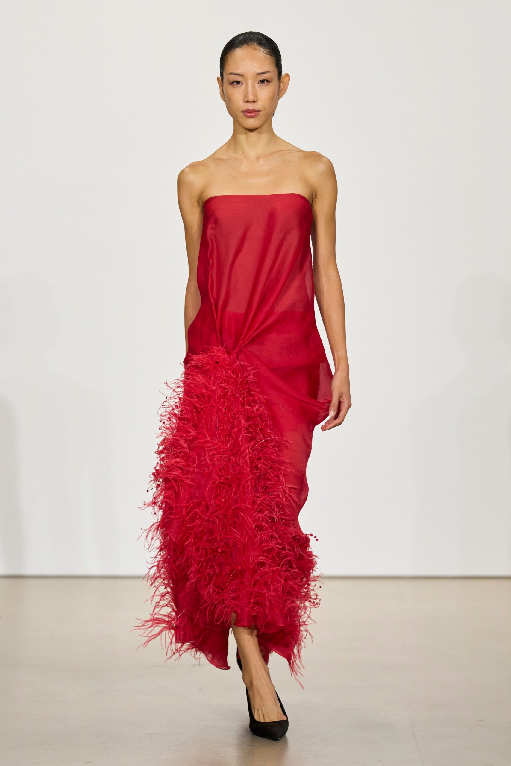 Sheer crimson column dress, ruched into a dense ostrich-feather burst at one hip. Black pointed pumps disappearing under the wavering hem. Crossing a hotel lobby after midnight with petals stuck to the carpet.