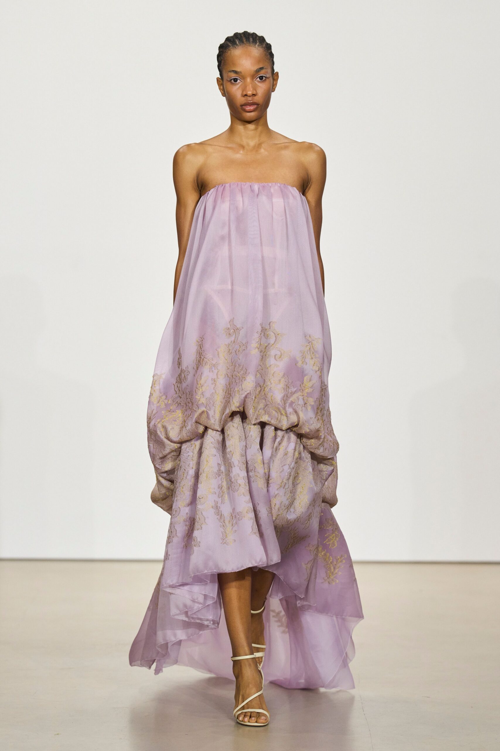 Strapless lilac organza gown, gold botanical brocade collapsing into a gathered bubble skirt. White barely-there straps crossing the toes. Garden wedding lawn after rain, hem drinking from the grass.