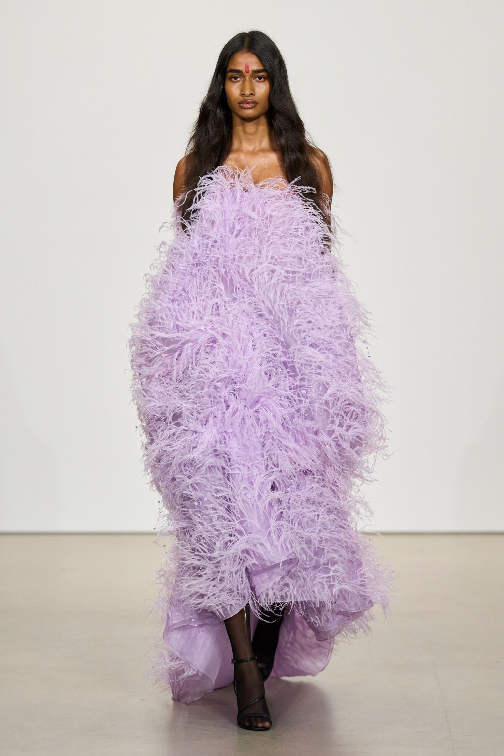 Strapless lilac gown buried in ostrich-feather fluff over sheer tulle. Black ankle-strap sandals disappearing under the dragging hem. Temple wedding reception, jasmine gone warm in the courtyard.