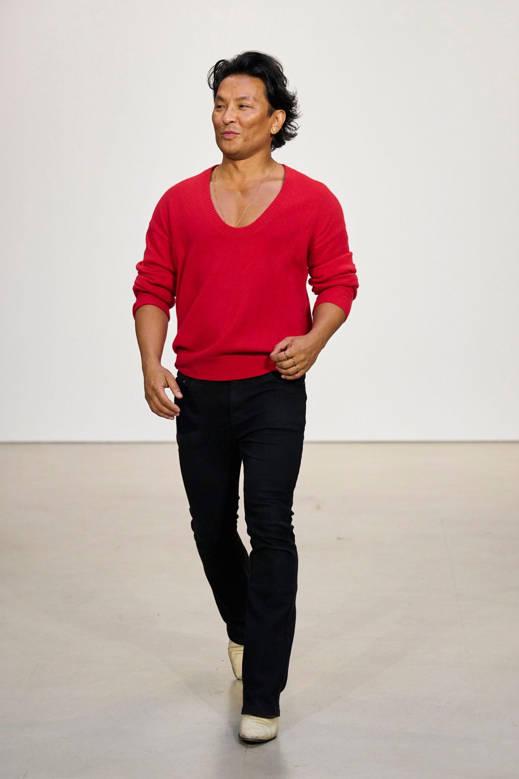 Scarlet fine-knit sweater with a deep scooped ribbed neckline. Thin gold chain disappearing into the collarbone dip. Black slim jeans, clean front and close through the thigh. Cream leather boots with squared toes peeking under the hem. Sunday gallery walkthrough, still wearing the rings from last night.